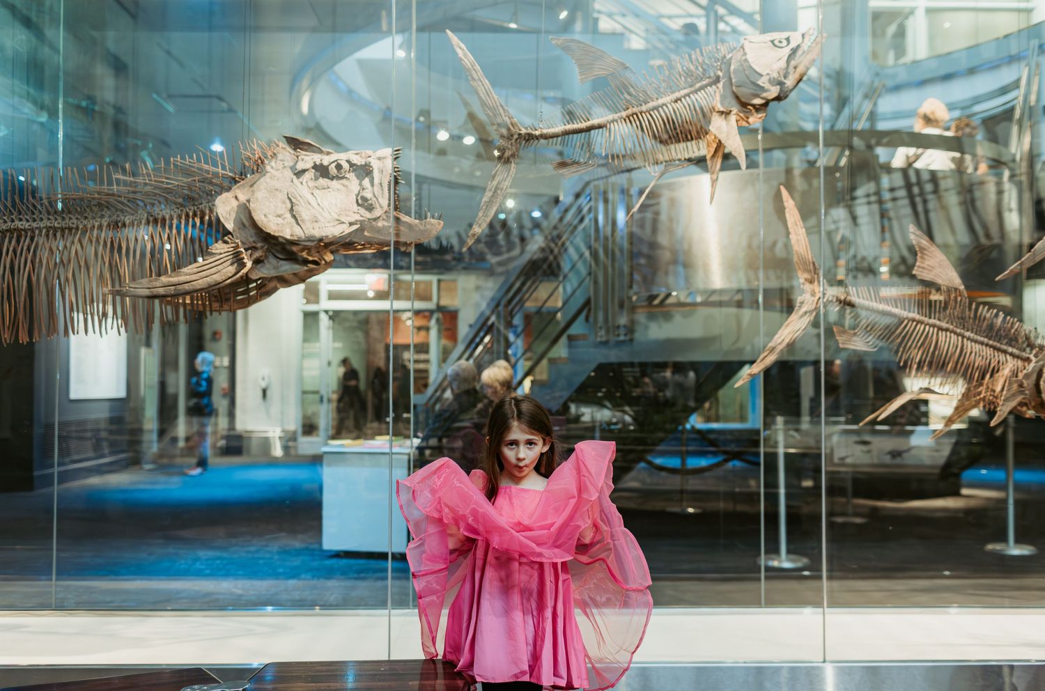 Child in pink dress poses in front of dinosaur skeleton display at natural history museum.