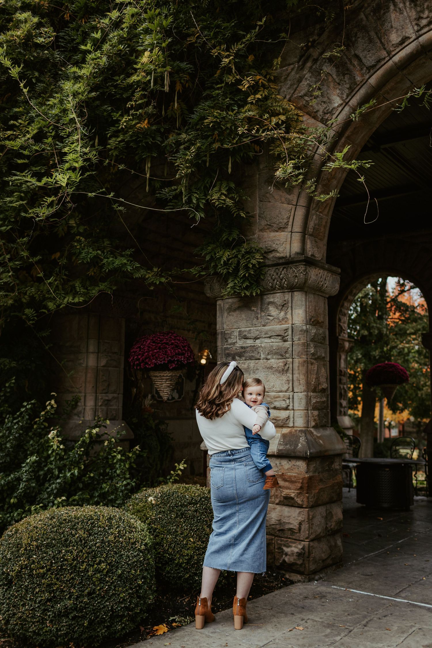 A parent holds a child while standing under a stone archway covered in lush green vines.