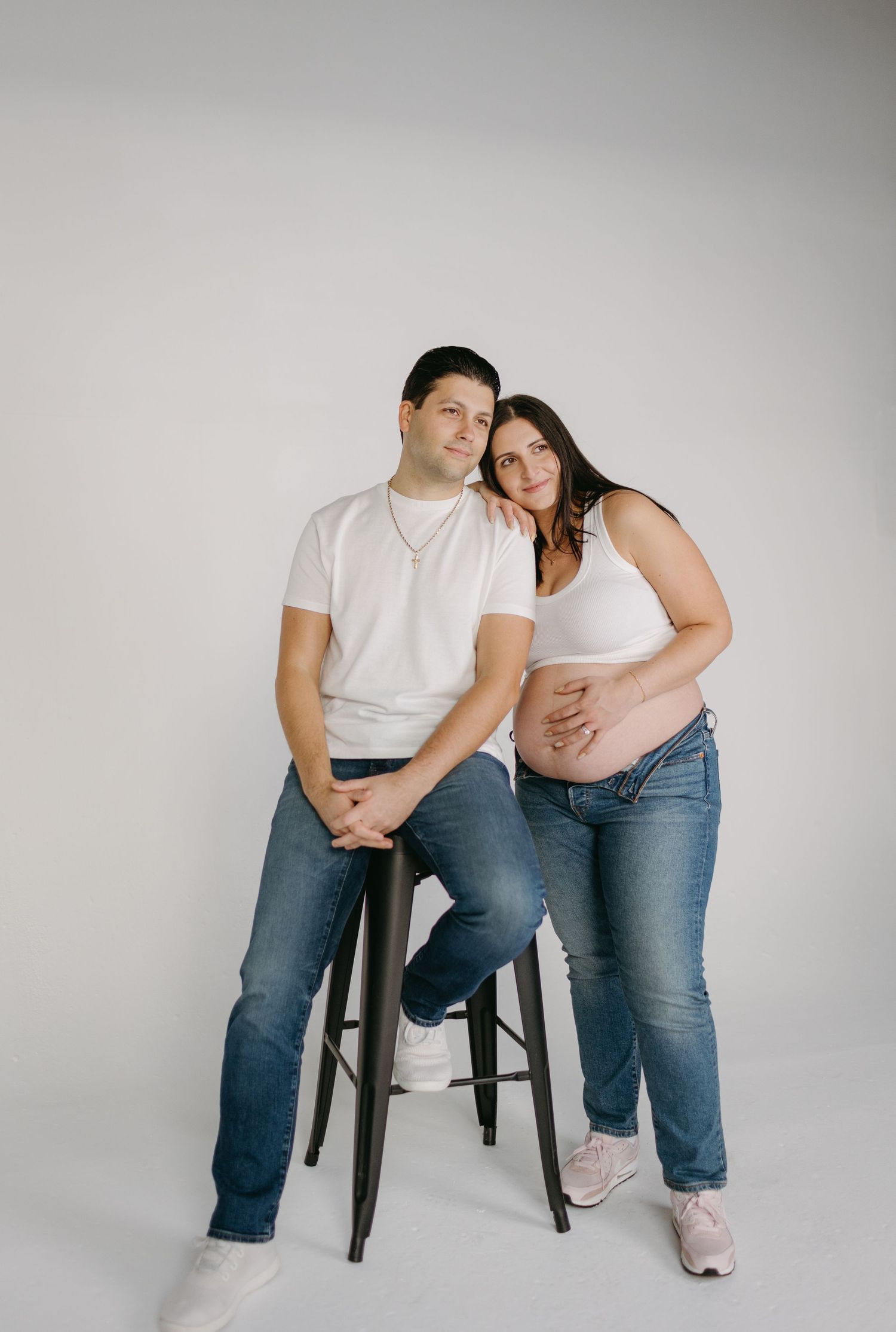 Maternity photo shoot featuring a couple in white tops and jeans posing together with a stool against a light backdrop.