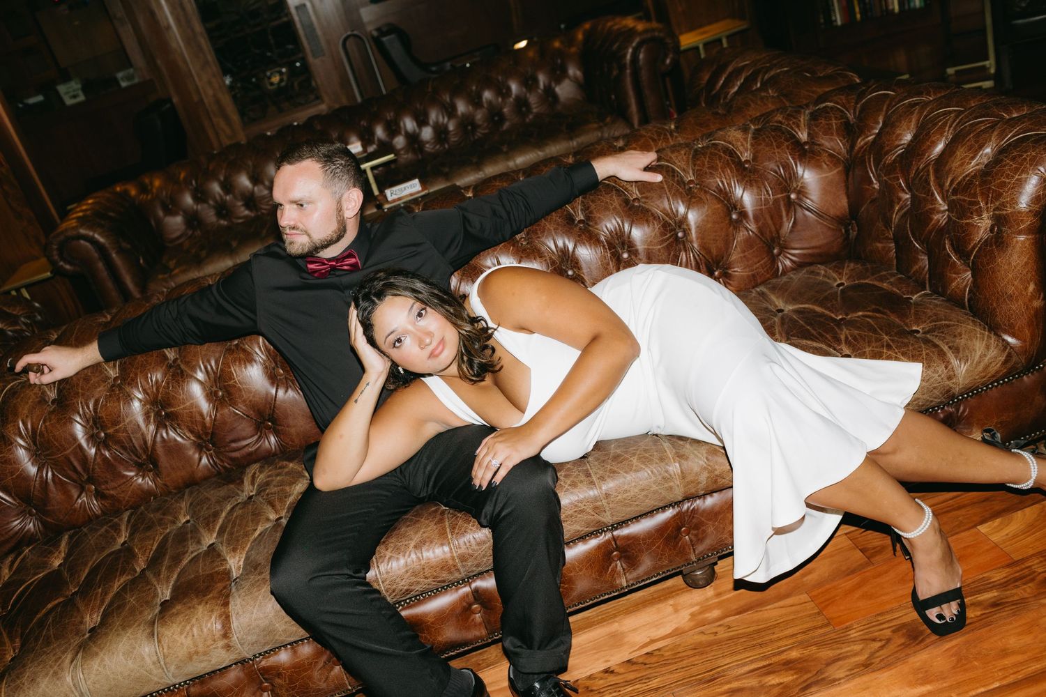 Couple relaxing on vintage brown leather Chesterfield sofa in dimly lit room.