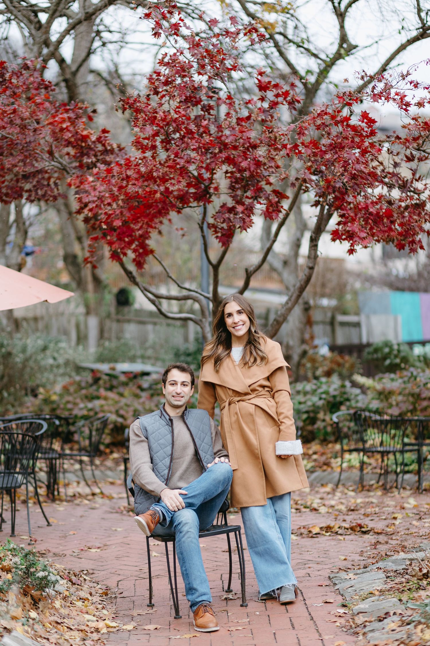 A couple poses beneath vibrant red autumn foliage on a brick pathway in a garden setting.