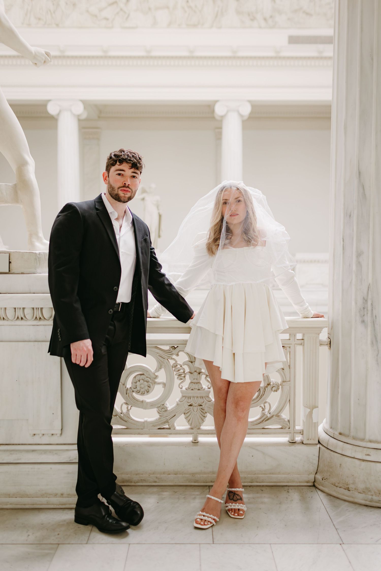 Stylish couple in formal attire poses in elegant white columned interior with ornate details.