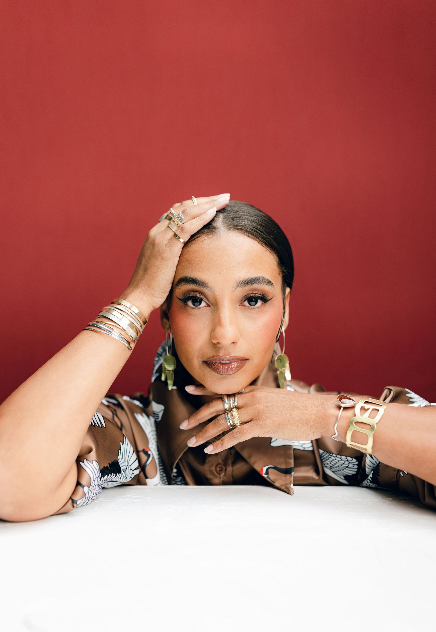 Portrait against a red wall featuring gold jewelry and bangles, with hands elegantly posed on a white surface.