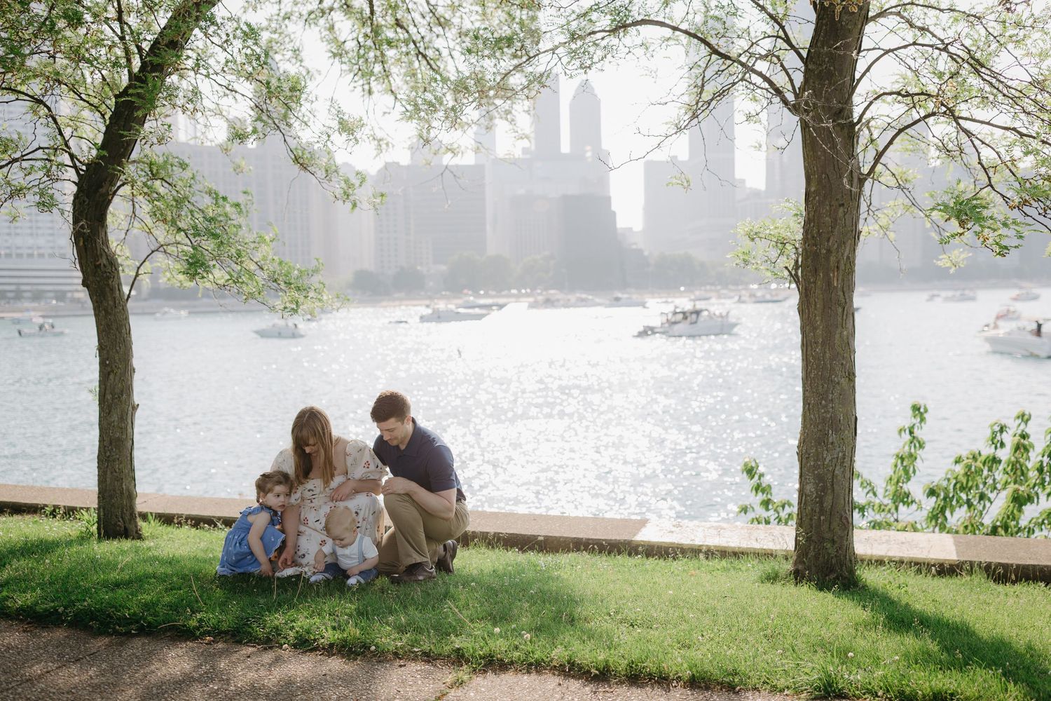 People enjoy a peaceful moment on grassy riverbank with city skyline and boats visible through trees on misty afternoon.