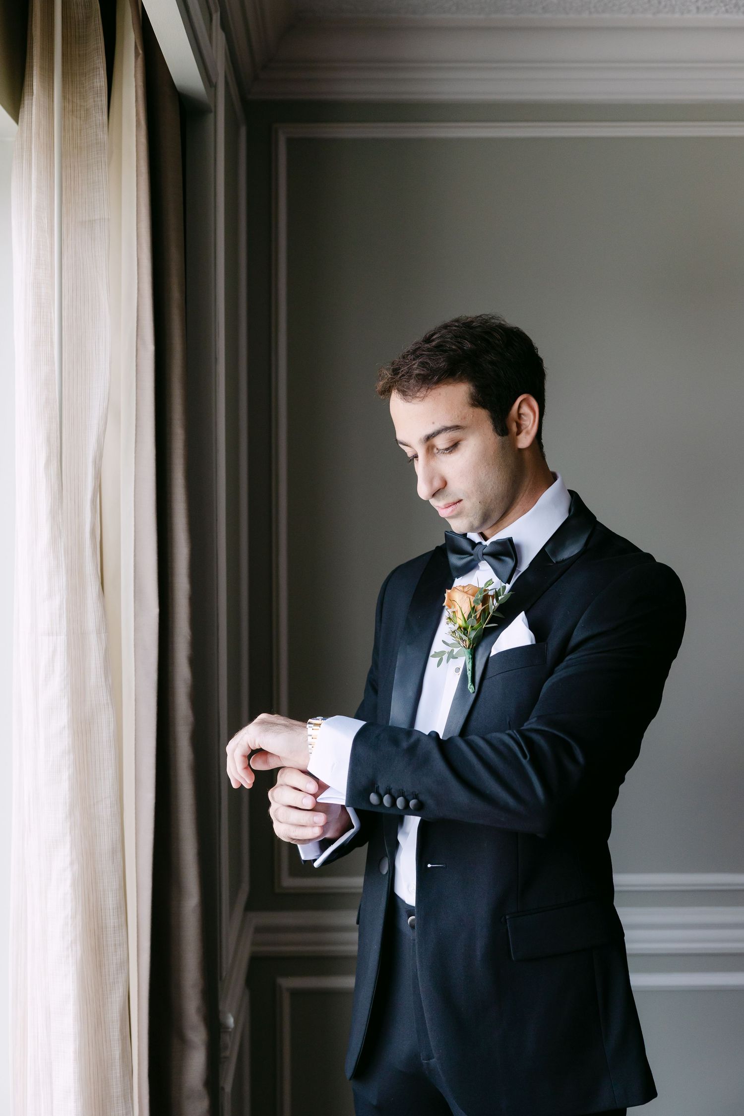 A person in a black suit adjusts their tie while standing near a window in an elegant room.