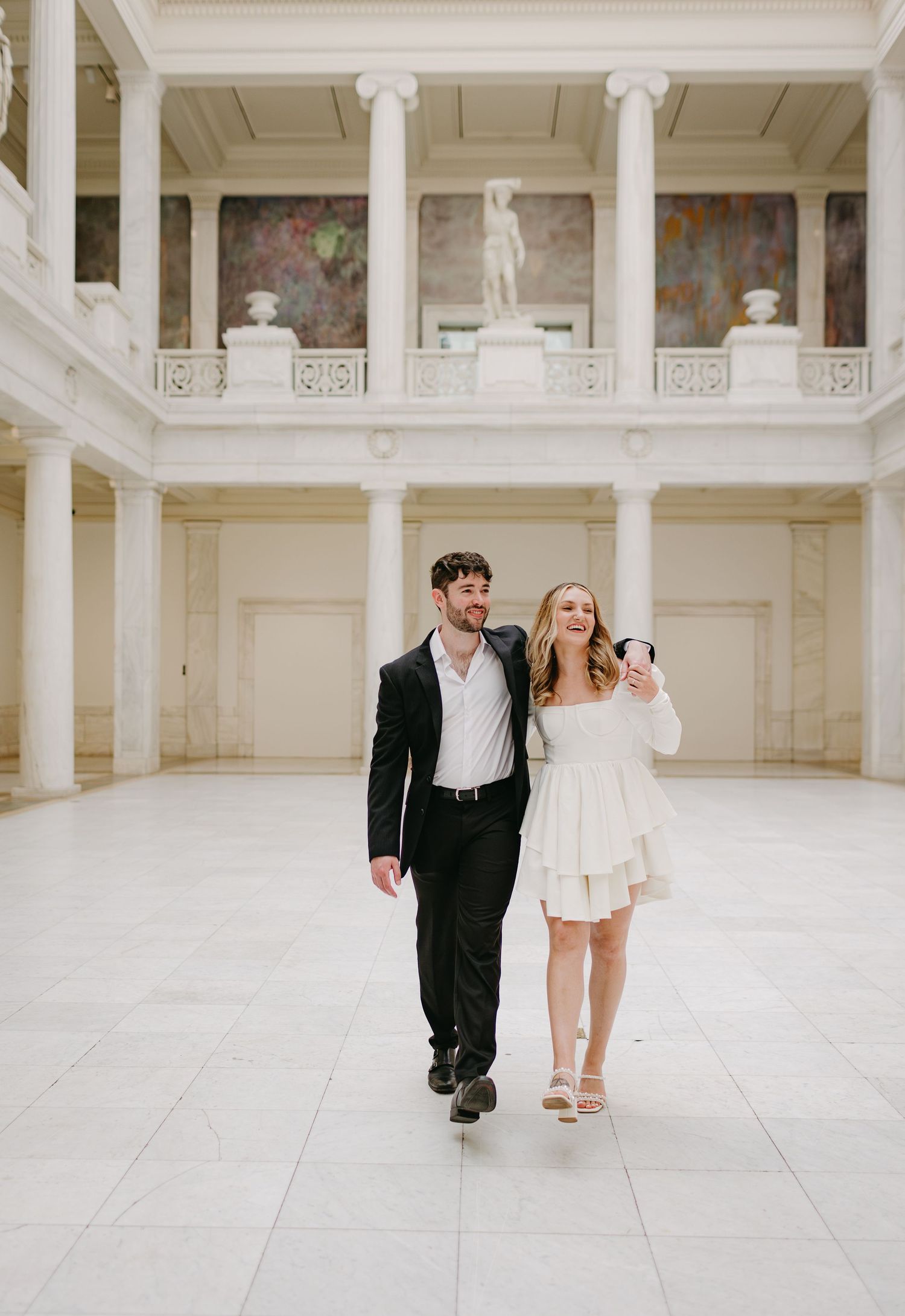 Couple walking through grand white marble hallway with classical architecture and artwork.