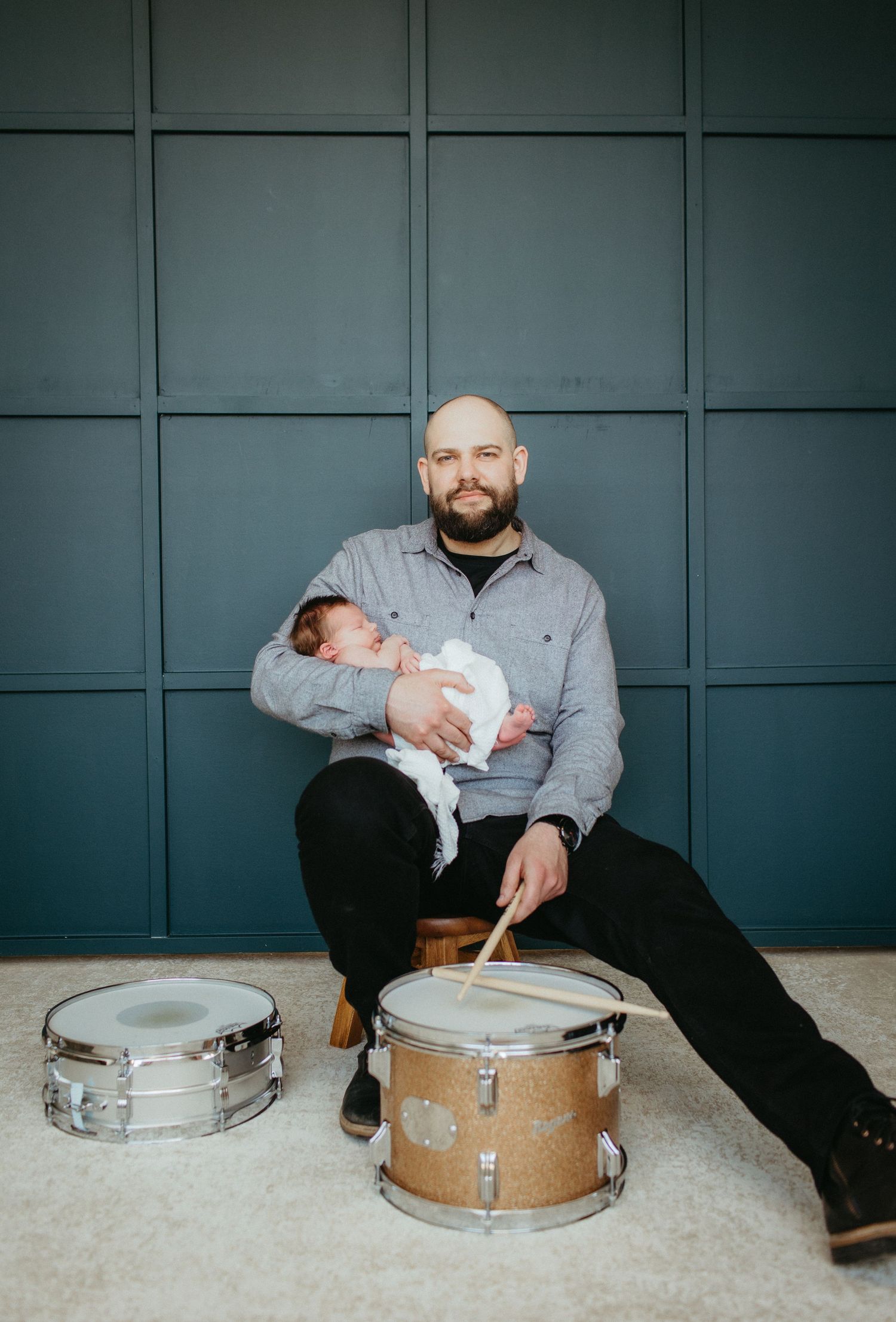 A drummer poses with a newborn baby against a dark paneled wall with two drums.
