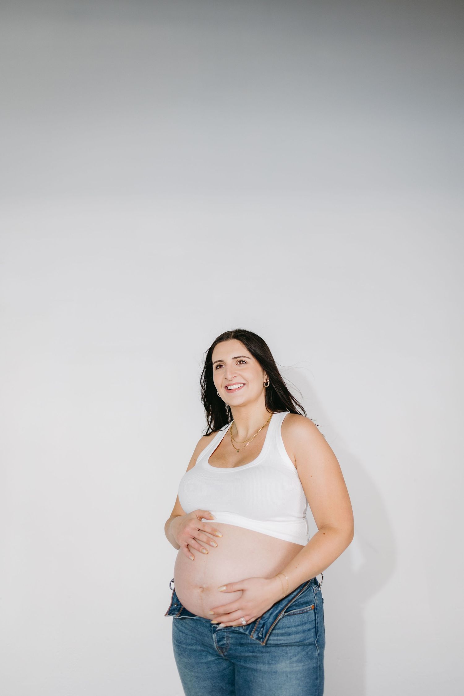 Pregnant woman in white tank top and jeans smiling against white wall during maternity photoshoot.