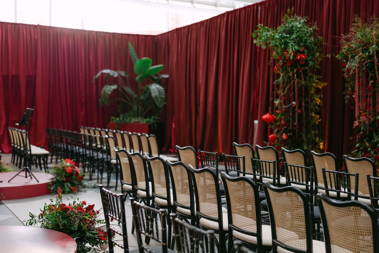 Empty rows of black metal chairs arranged in front of a red barn wall with green plants along the perimeter.