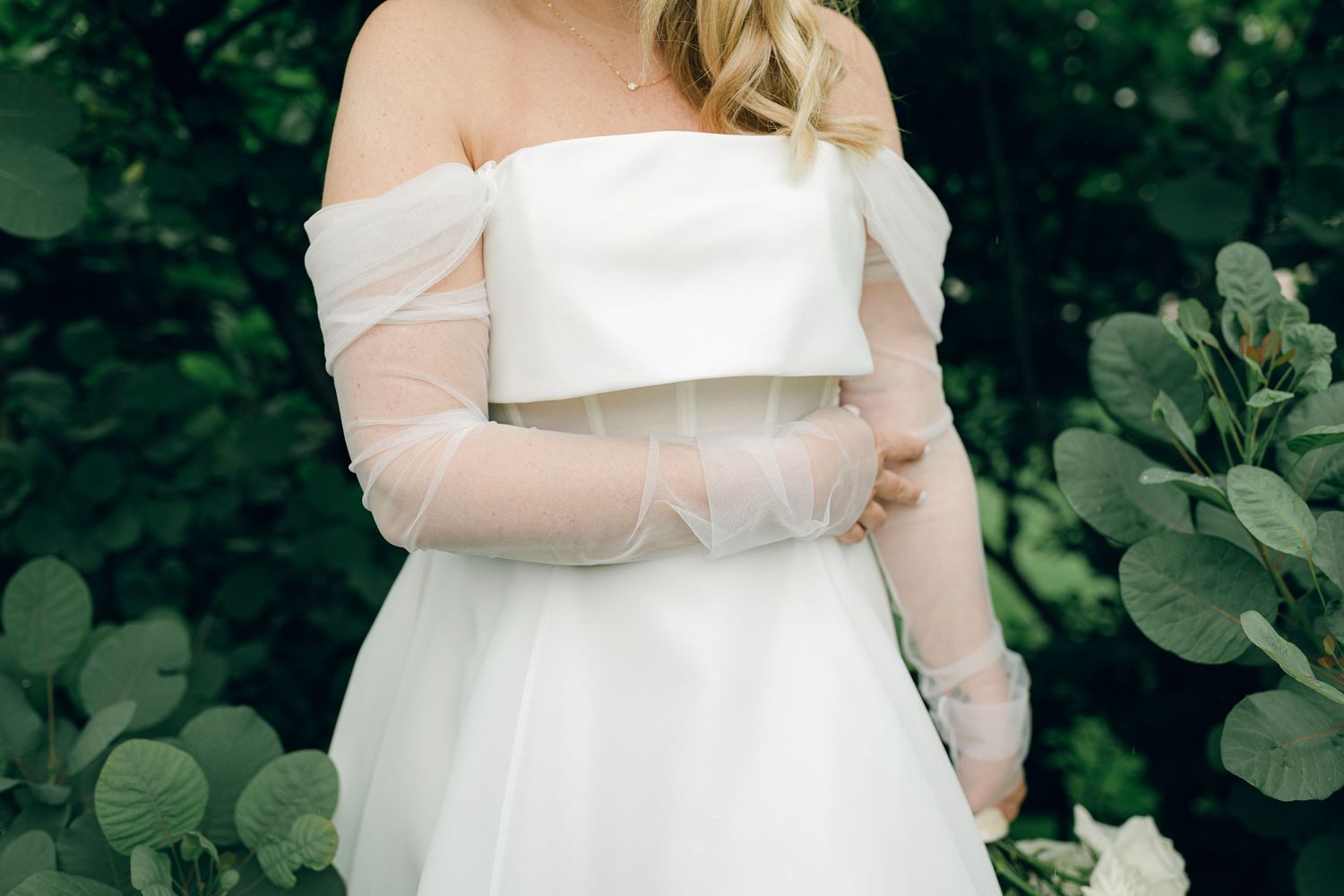 Detail of an off-shoulder white wedding dress with sheer long sleeves against green foliage background.