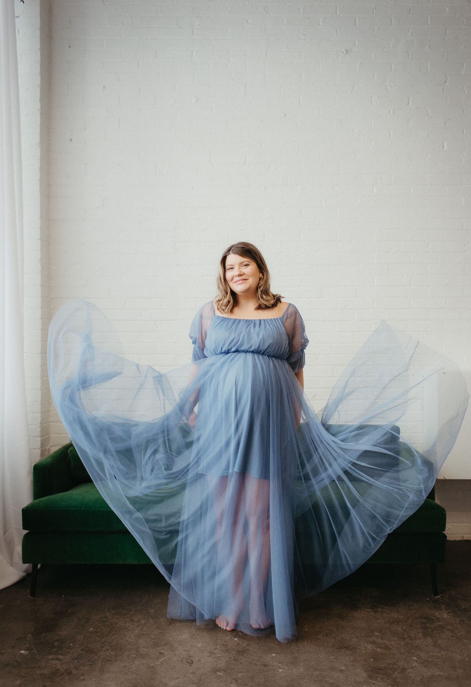 A flowing blue tulle maternity gown creates dramatic movement against a white wall and green velvet couch.