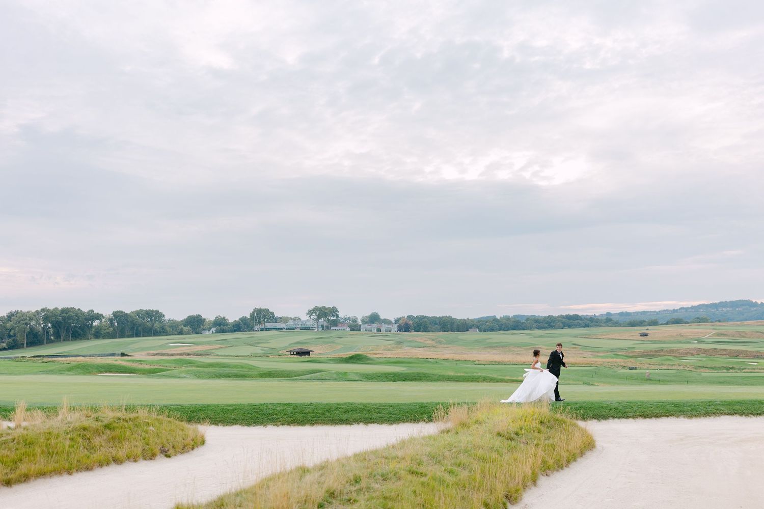 Bride and groom share a romantic moment on a winding golf course path surrounded by lush green fairways under a cloudy sky.