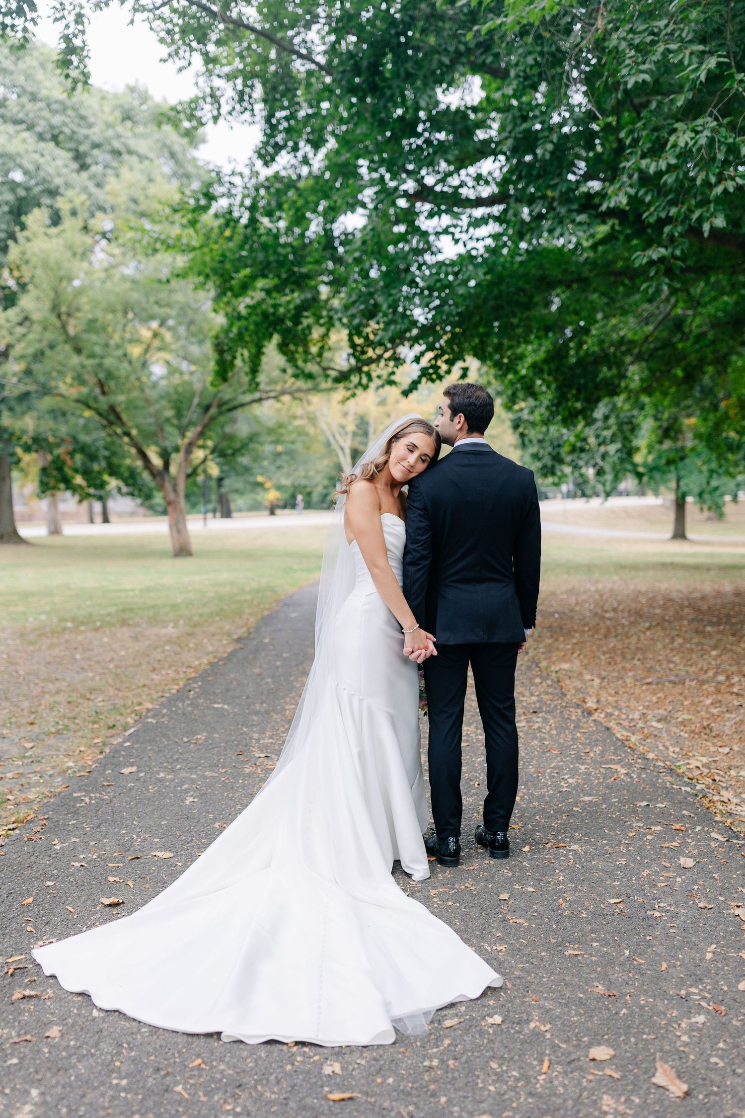 A couple in wedding attire walks along a tree-lined path with a flowing white bridal gown trailing behind.