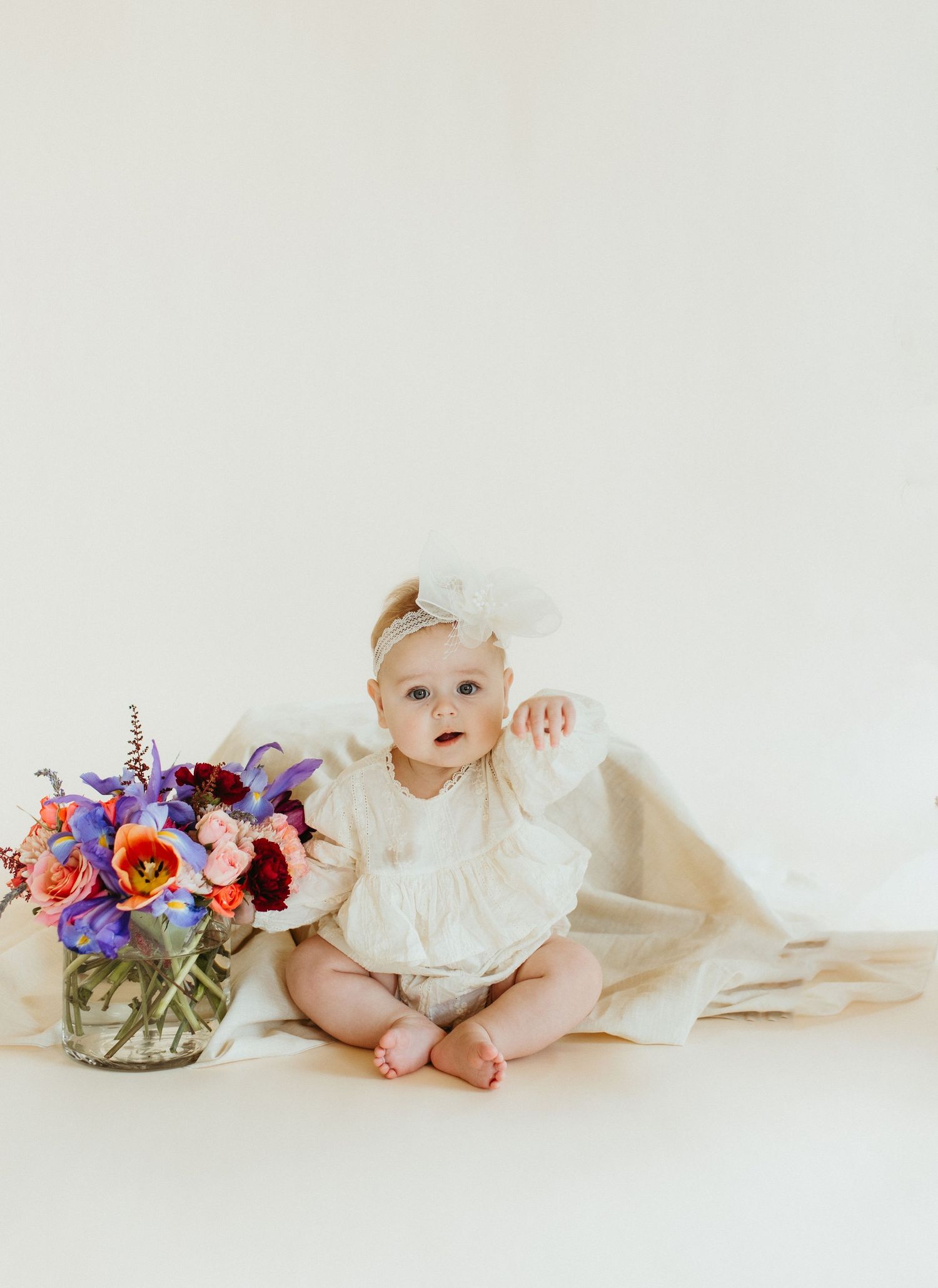 Baby in white lace dress sits next to colorful floral arrangement on light background.
