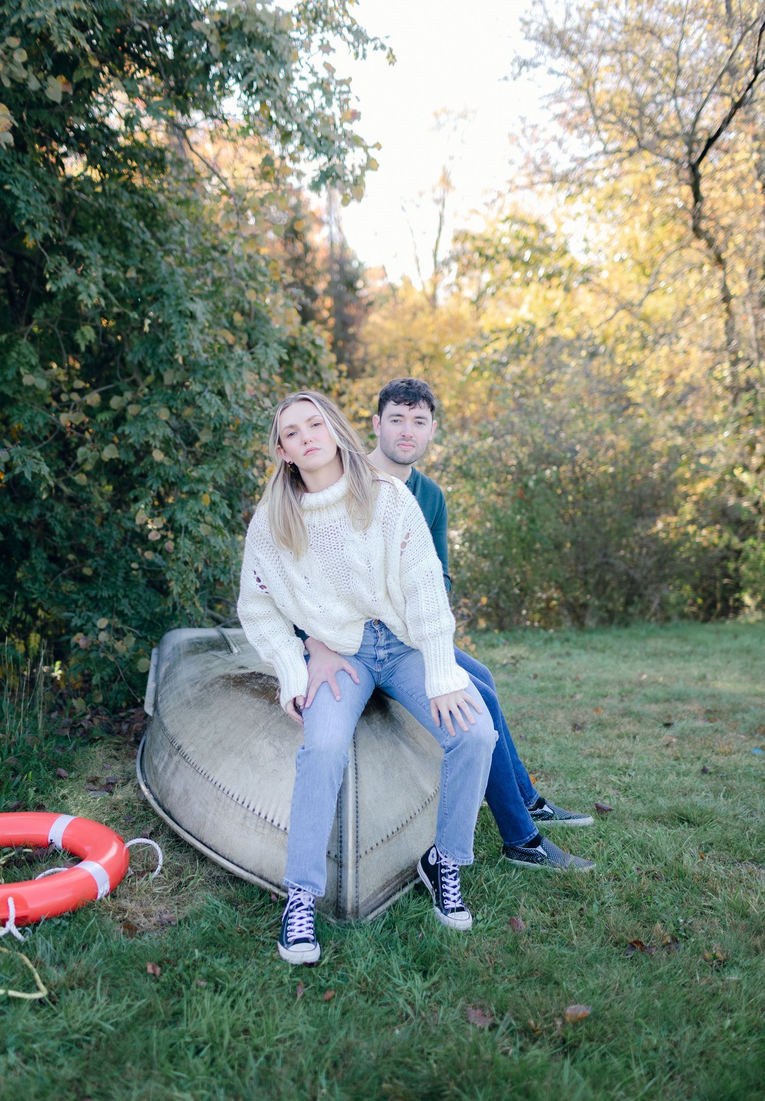 A couple sits together on an overturned boat surrounded by autumn foliage in a peaceful outdoor setting.