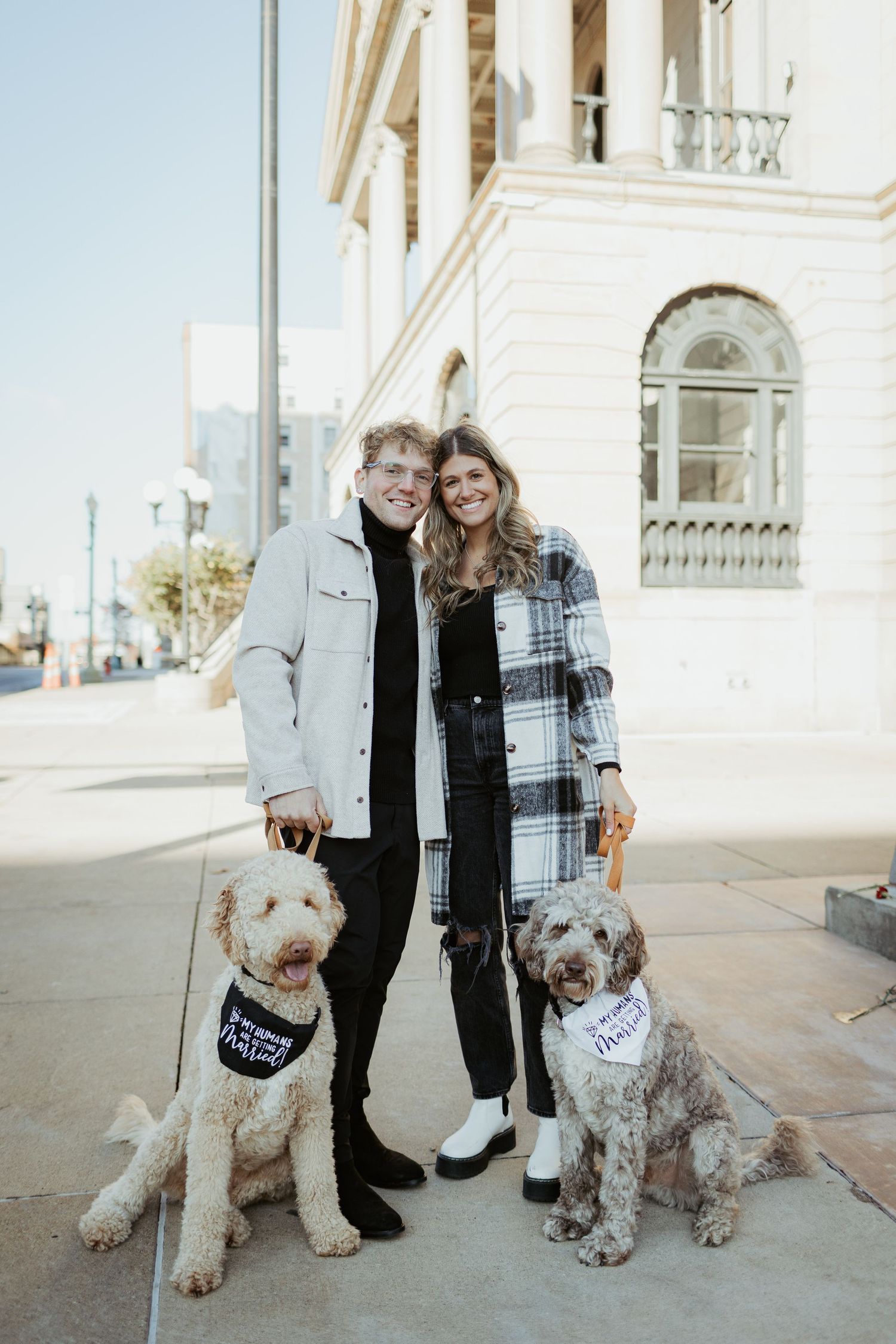 Couple posing with two golden doodle dogs in front of a white building on a sunny day.