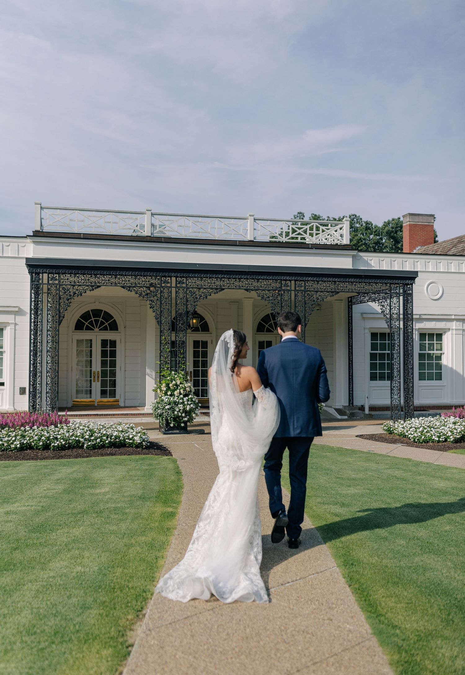 Elegant couple walking down garden path toward white mansion with arched porch and beautiful landscaping.