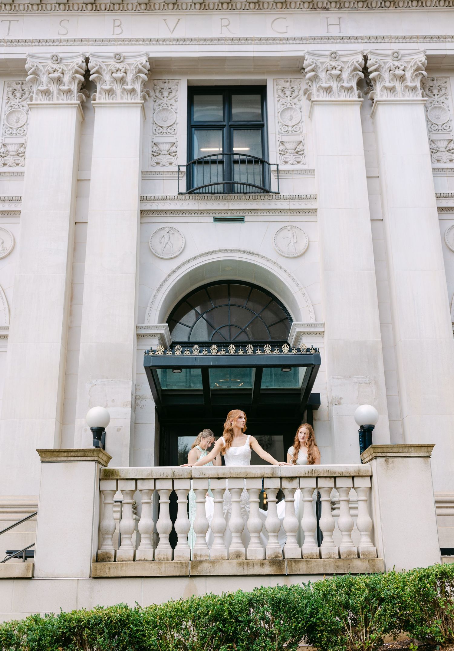 Bride and bridesmaids stand on ornate white balcony of elegant neoclassical building with arched windows.