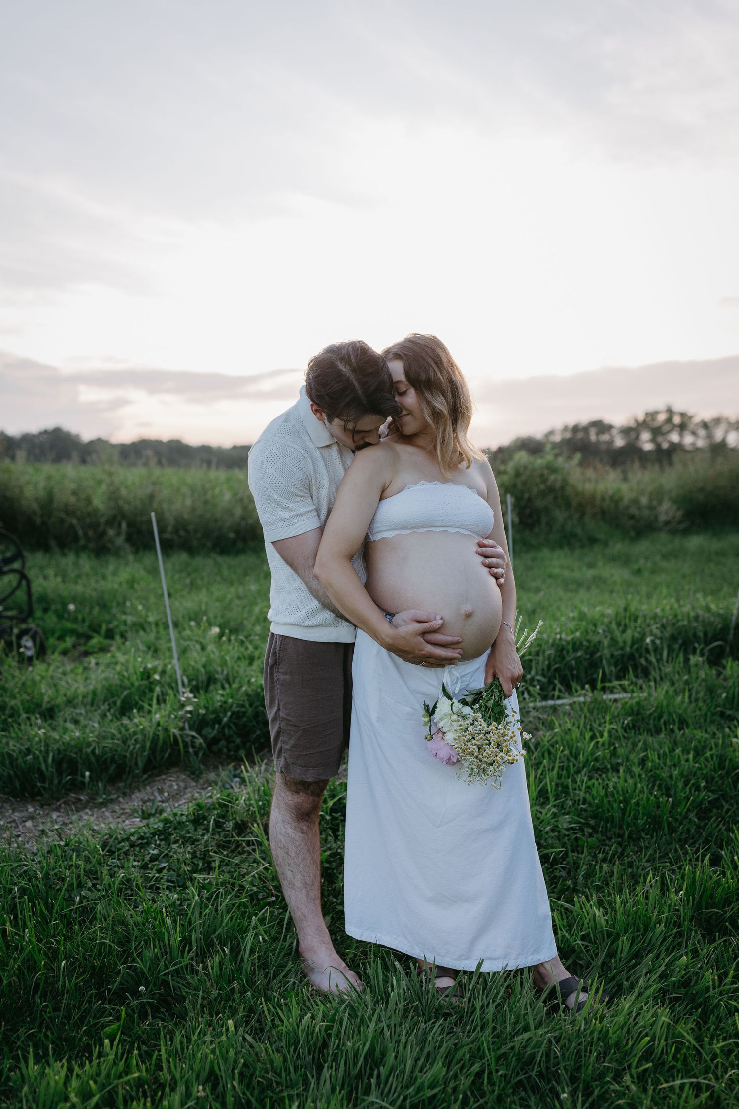 Intimate maternity photo series showing couple embracing in green summer field at dusk.