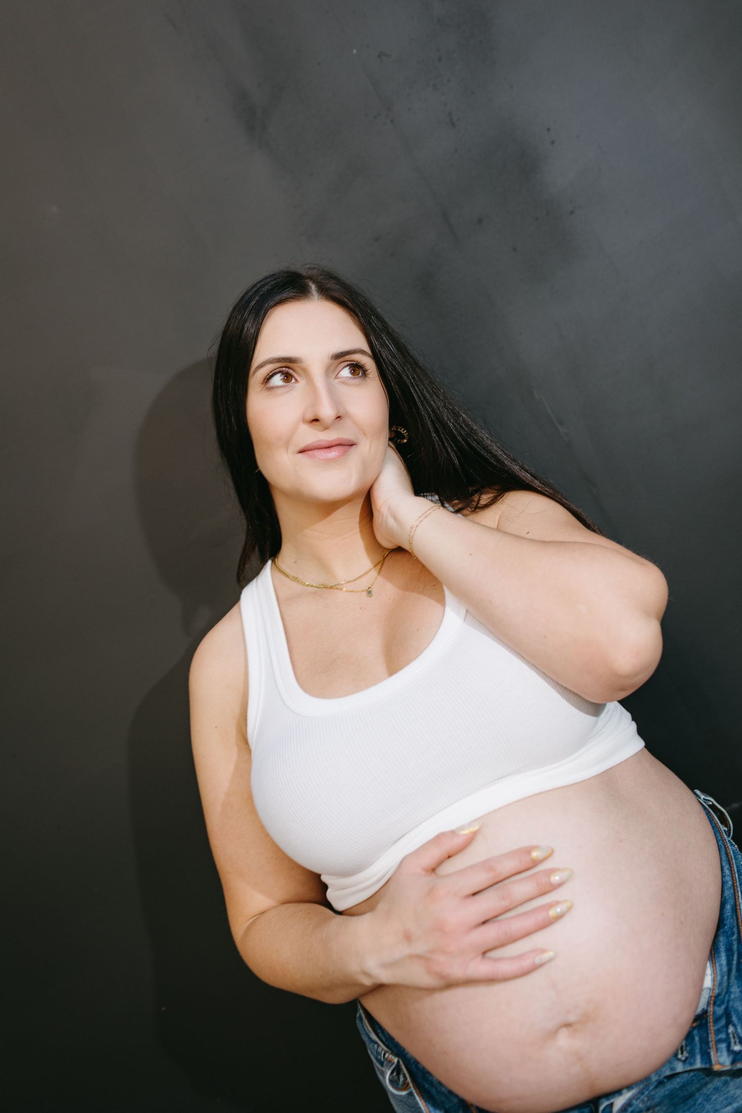 Pregnant woman in white crop top poses against dark wall while touching her neck.