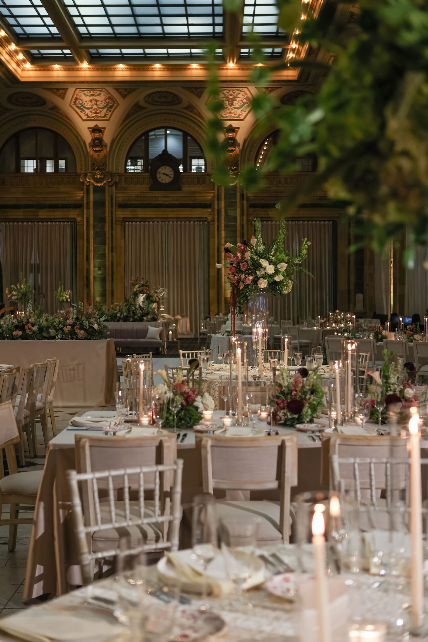 Elegant wedding reception in grand ballroom with candlelit tables, floral centerpieces, and ornate arched windows.