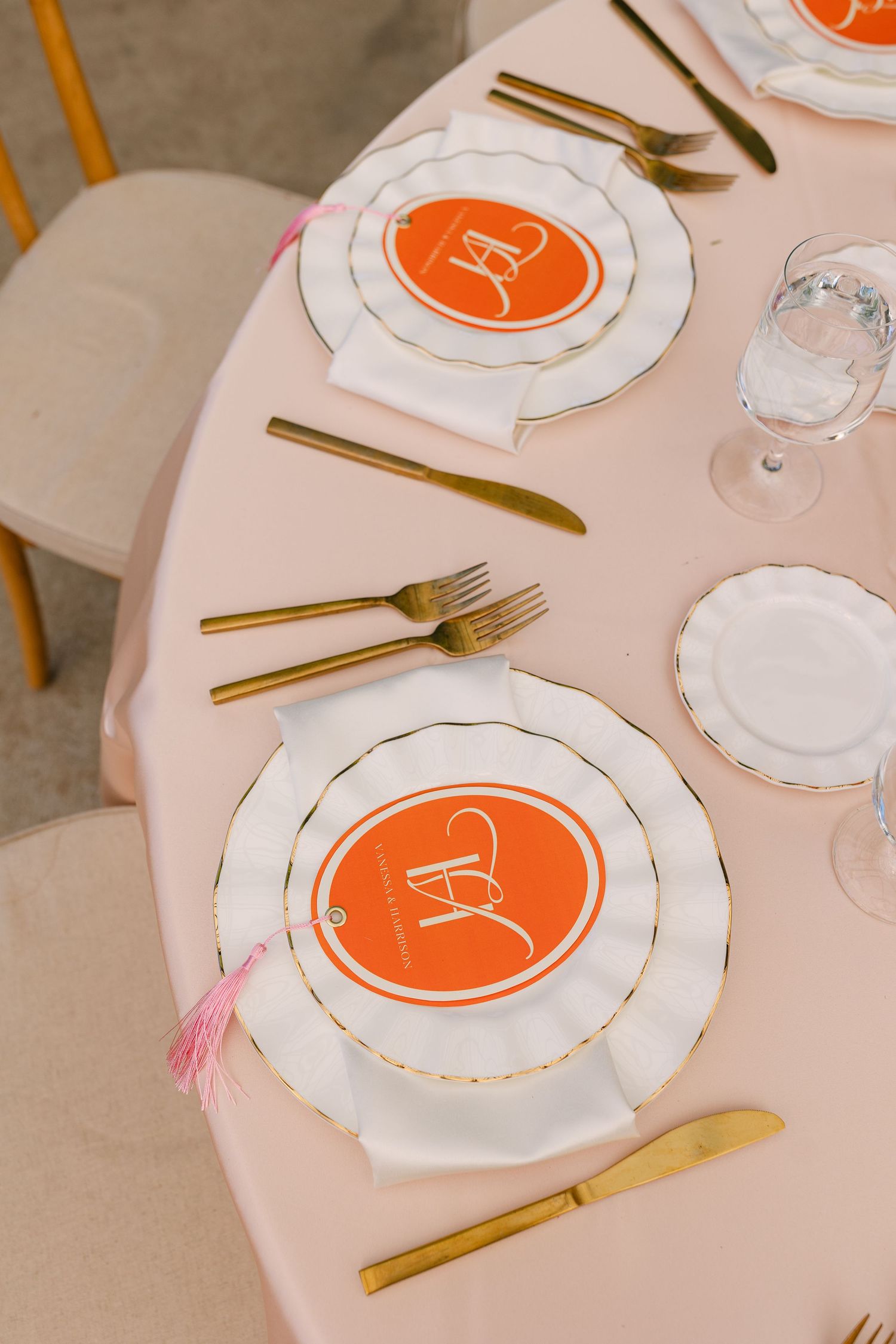 Elegant wedding table setting with orange monogram plates, gold cutlery, pink tassel napkins on blush tablecloth.