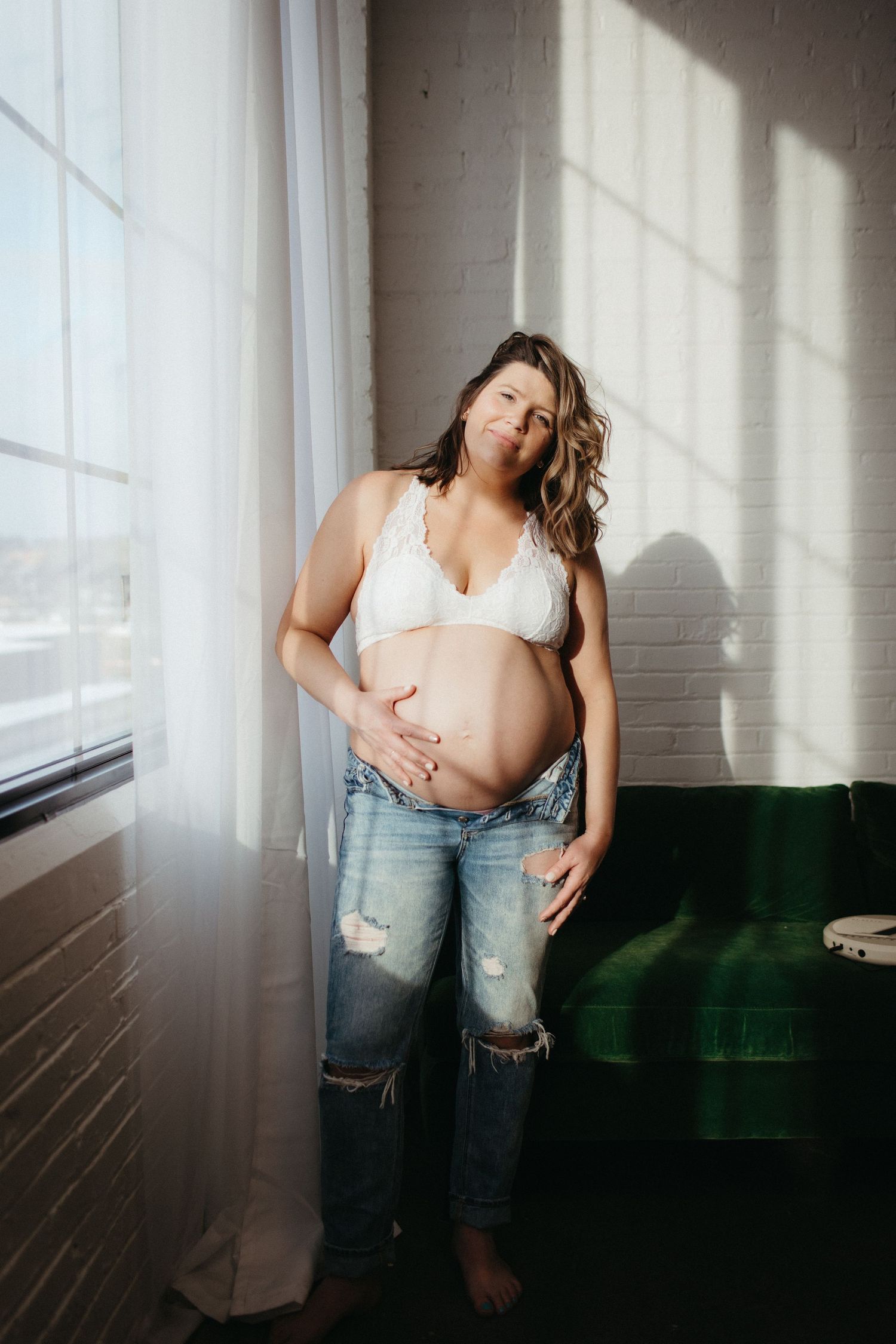Natural window light illuminates a maternity portrait with green velvet couch and distressed denim.