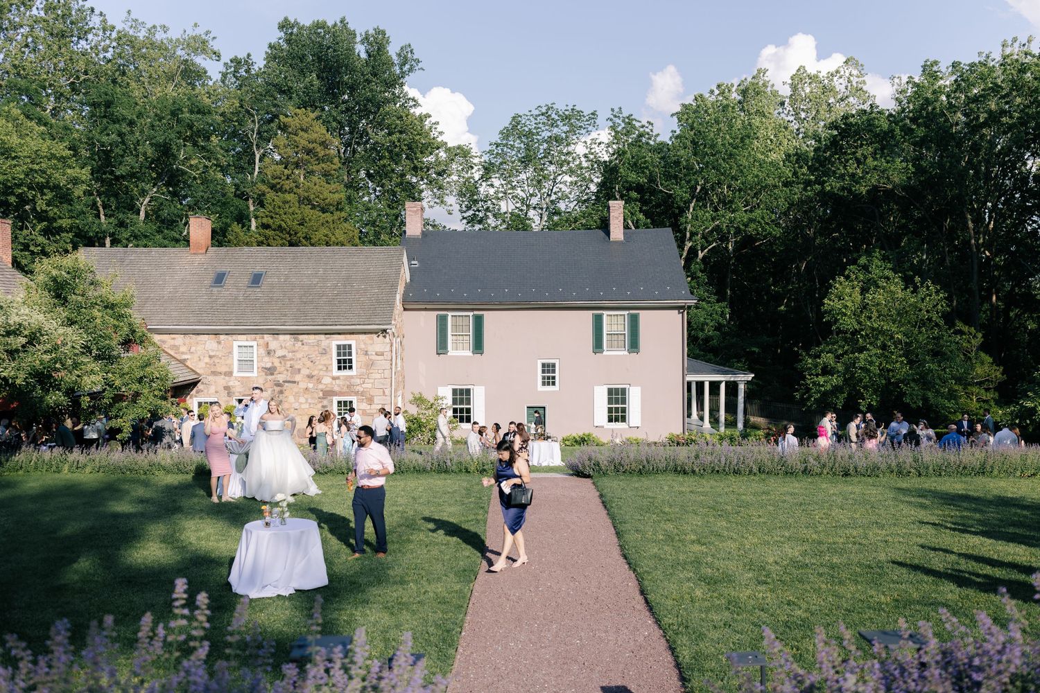 Guests gather on a brick path leading to a historic stone house with gardens during a special outdoor event.