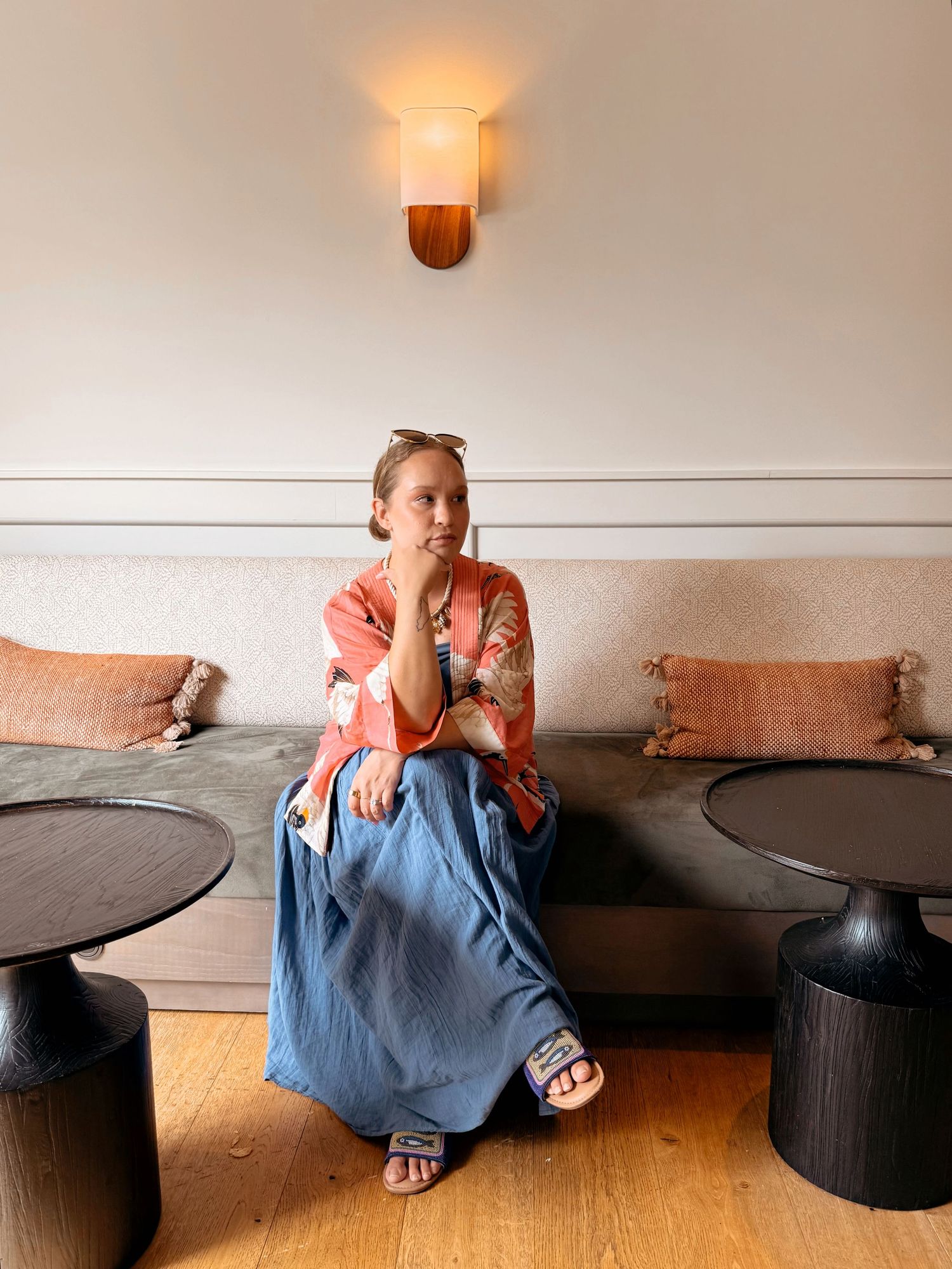 Woman in coral top and blue skirt sits on gray bench beside dark side tables in a warmly lit lounge.