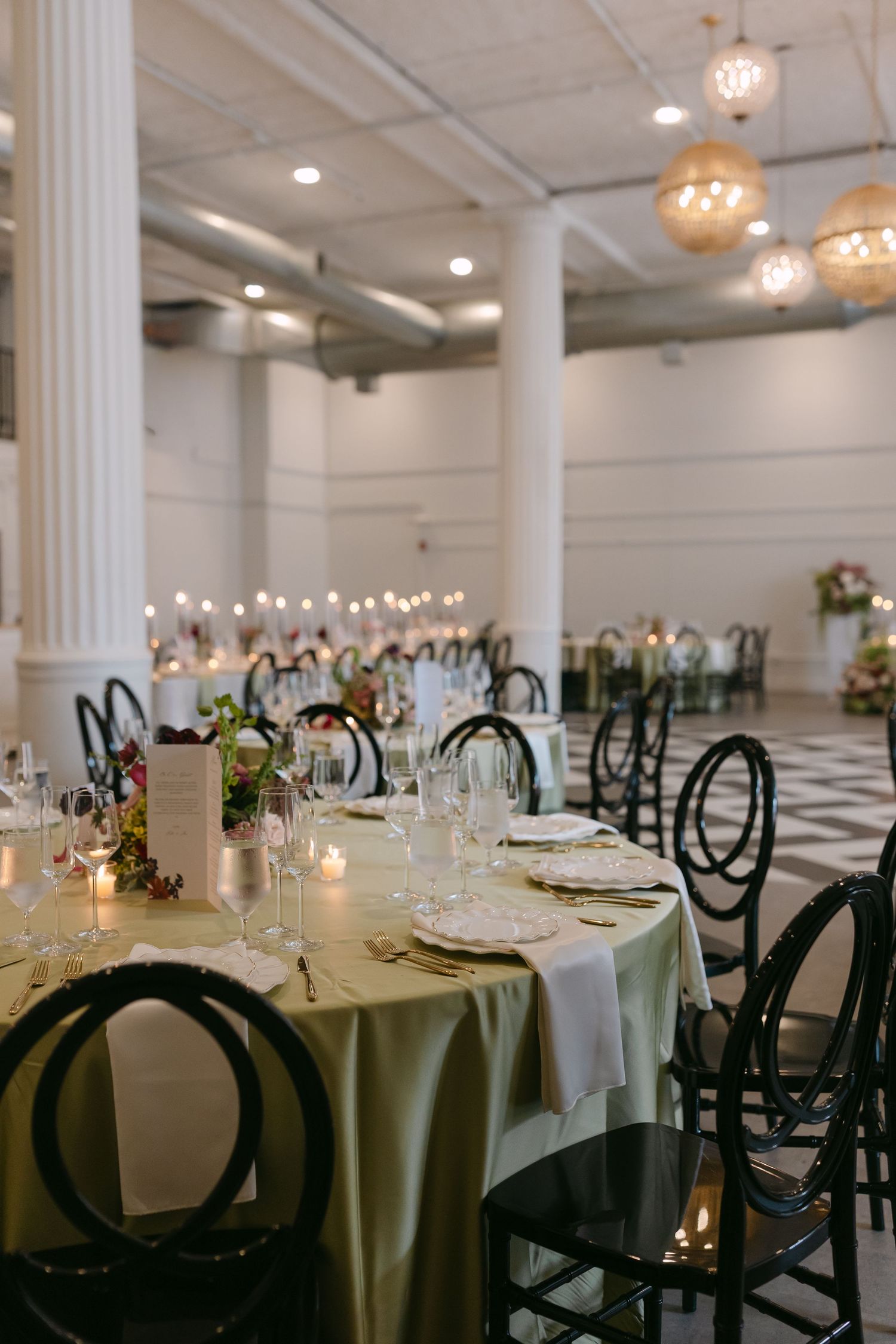 Elegant wedding reception hall with sage green tablecloths, black circular chairs, candles, and woven pendant lights.