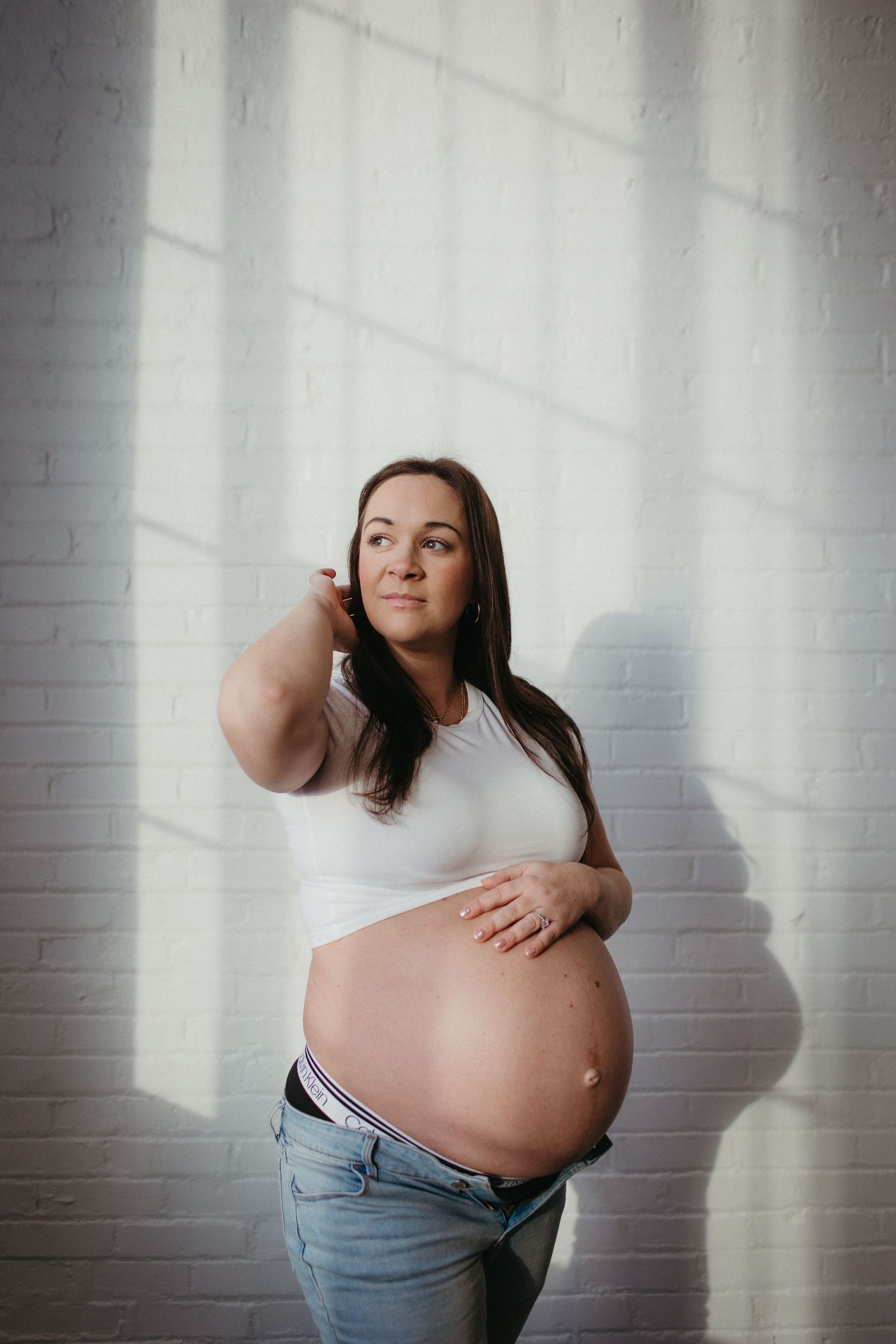 Maternity photo of an expectant mother in a white top and jeans against a white brick wall.