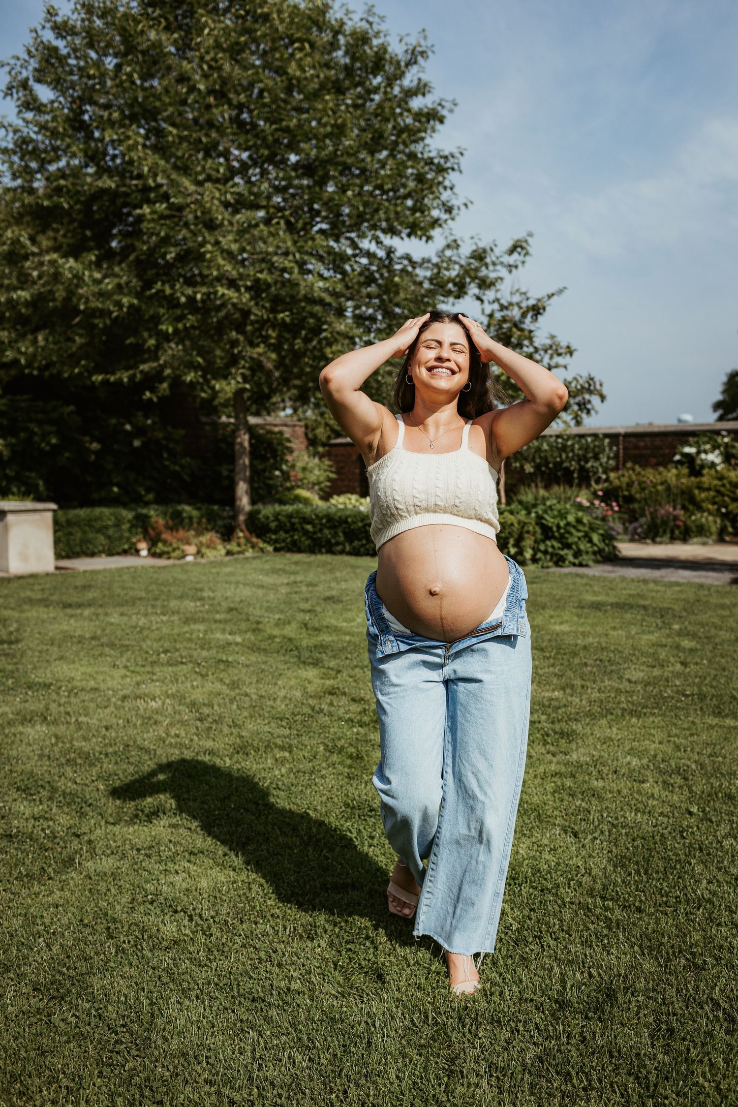 An expectant person stands on grass wearing jeans and crop top with hands raised joyfully.