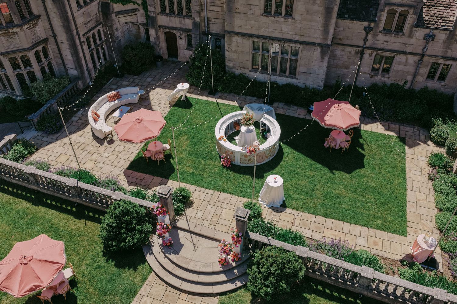Aerial view of an elegant hotel courtyard garden with fountain, pink umbrellas, seating areas, and manicured lawn.