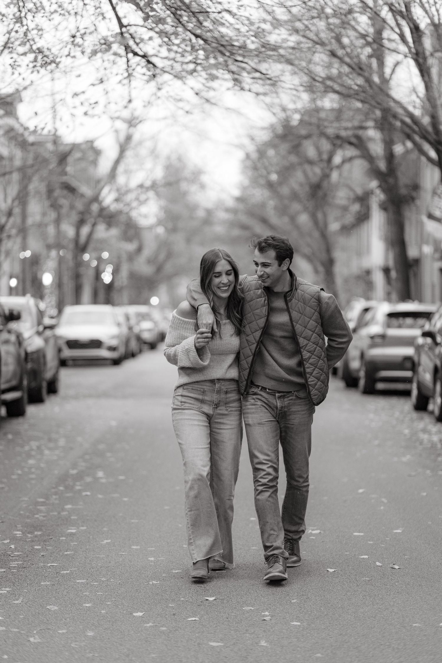 A couple walks together laughing on a tree-lined street with parked cars.