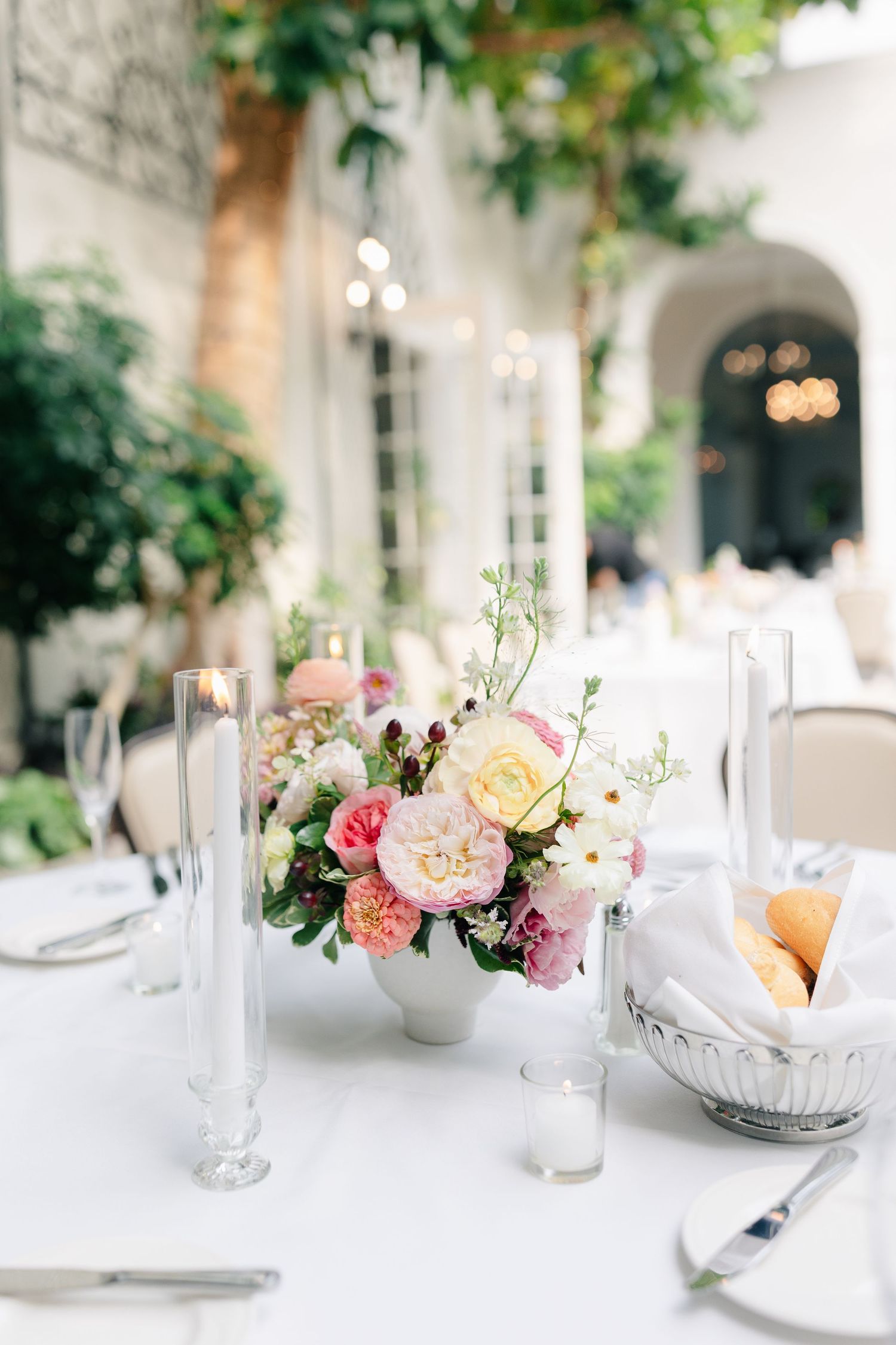 Elegant wedding reception table settings with soft pink and peach floral centerpieces and white candles in a bright venue.