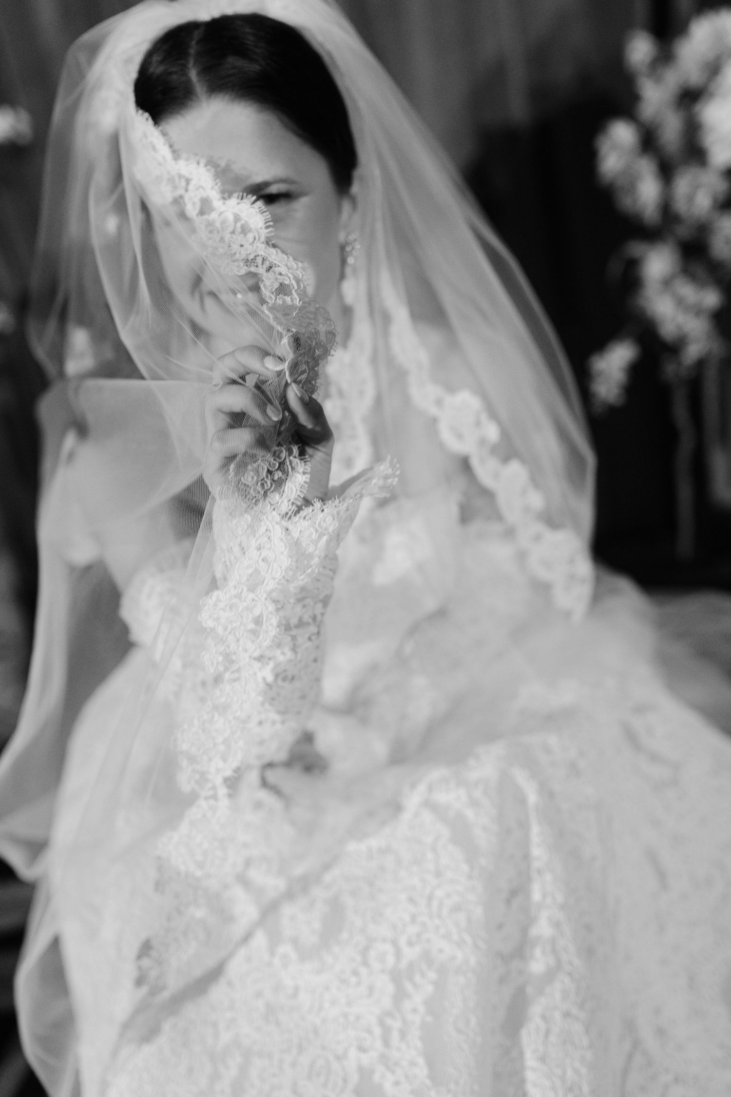 Black and white artistic shot of a bride in lace veil and dress holding delicate florals near her face.