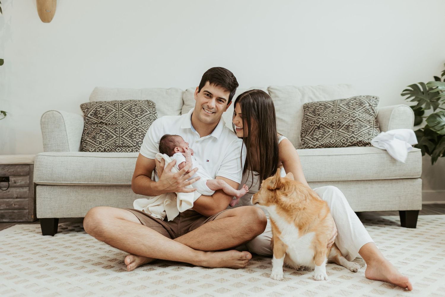 A family sits on floor with their newborn baby and dog in a cozy living room setting.
