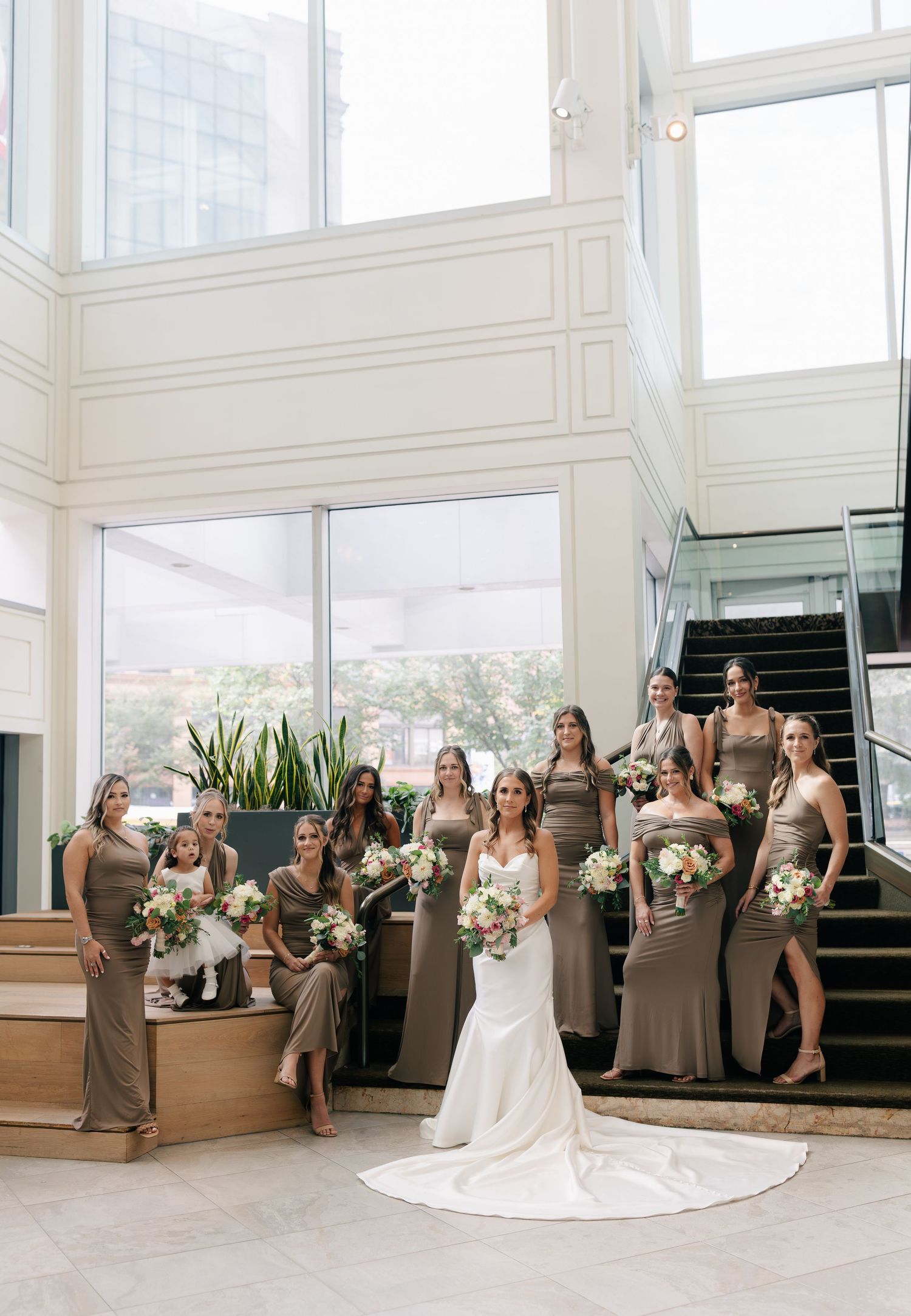 Wedding party in neutral taupe dresses poses elegantly in a bright modern space with large windows.