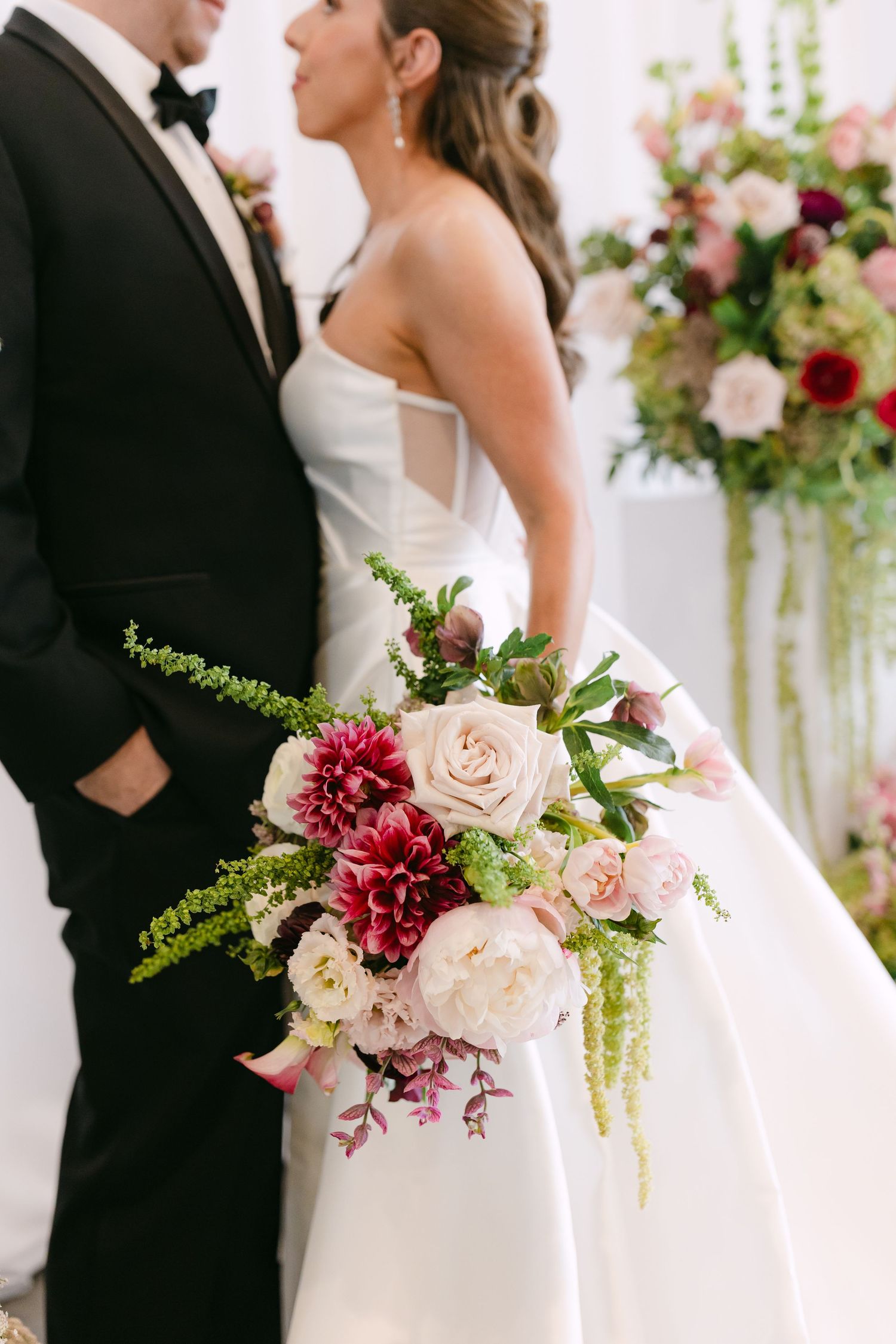 Bride holding lush bouquet of pink peonies, dahlias, and greenery while standing with groom in black tuxedo.