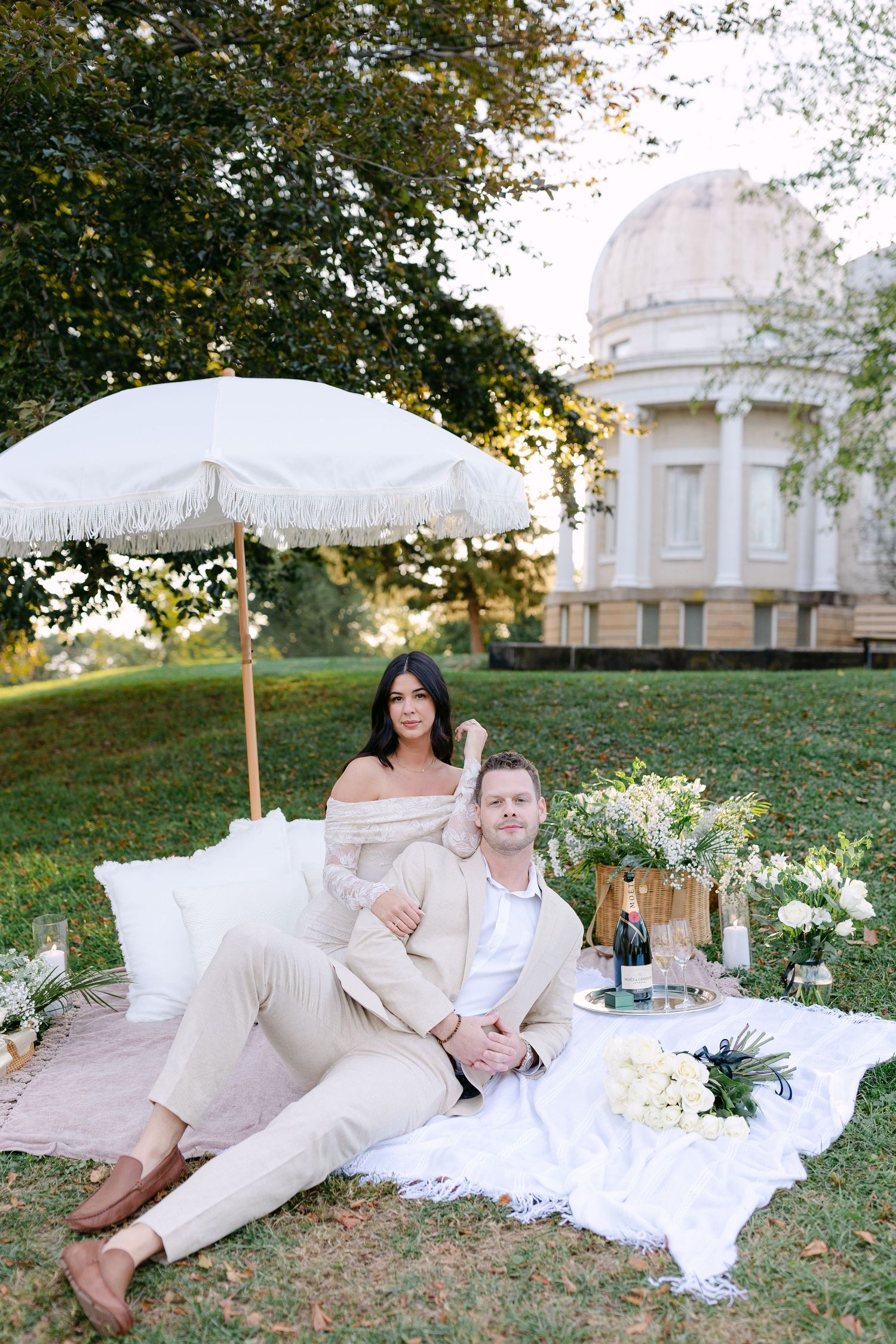 A romantic picnic setup with a white parasol and blanket on grass near a domed building at sunset.