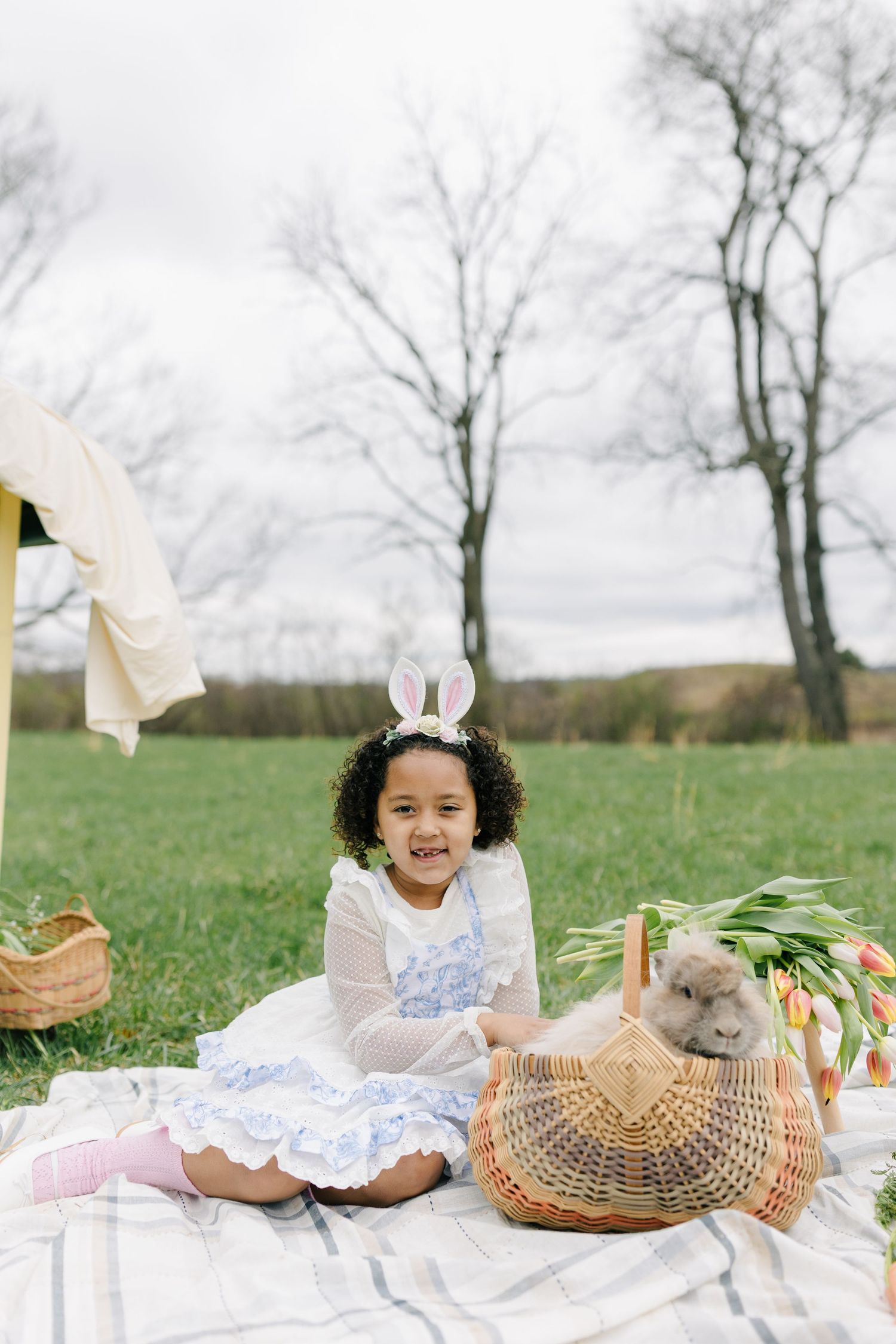 A charming spring picnic setup with bunny ears, a wicker basket, and white lace blanket spread on green grass.