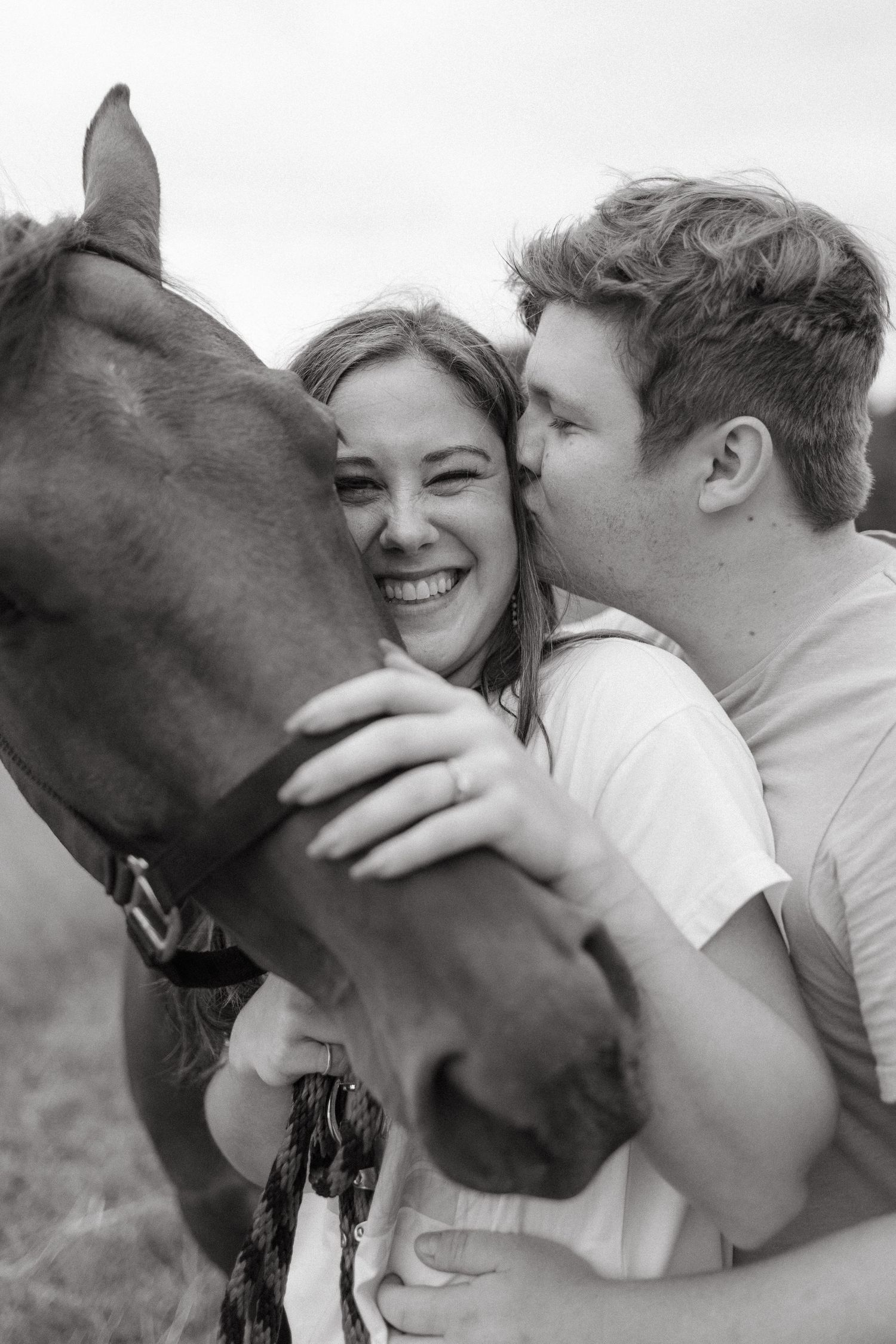 A joyful moment shared between a couple and a brown horse in a black and white photograph.