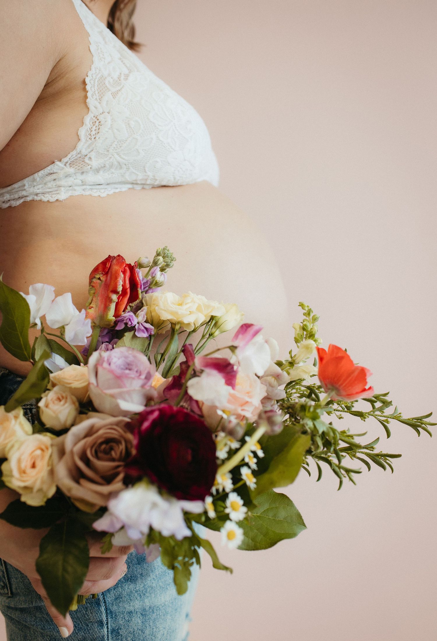 A colorful bouquet of roses, peonies and mixed flowers held against a person wearing white lace and denim.