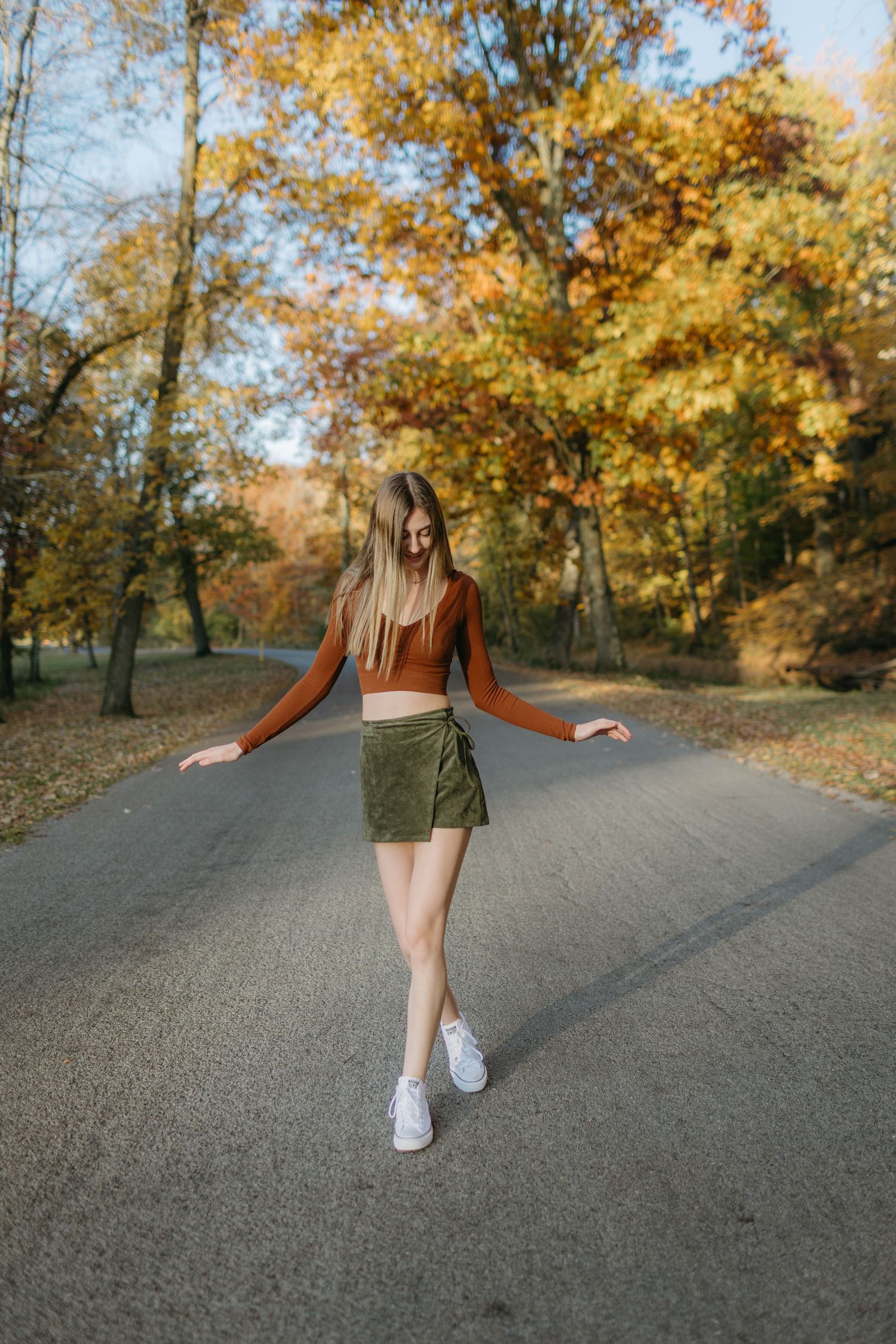 Person in rust sweater and olive skirt dancing on autumn road surrounded by golden trees.
