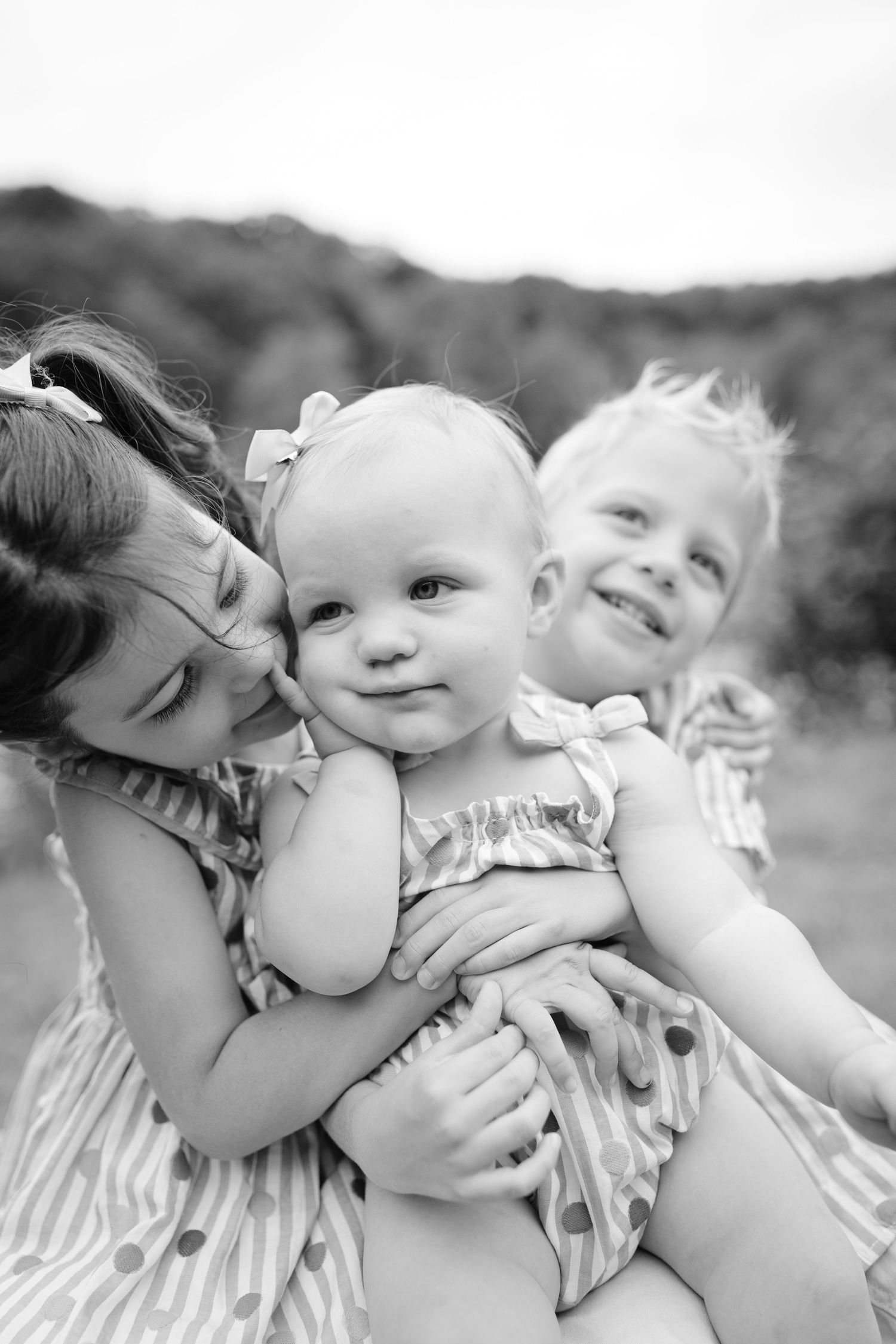 Black and white portrait of three siblings in matching dresses enjoying a sweet outdoor moment together.