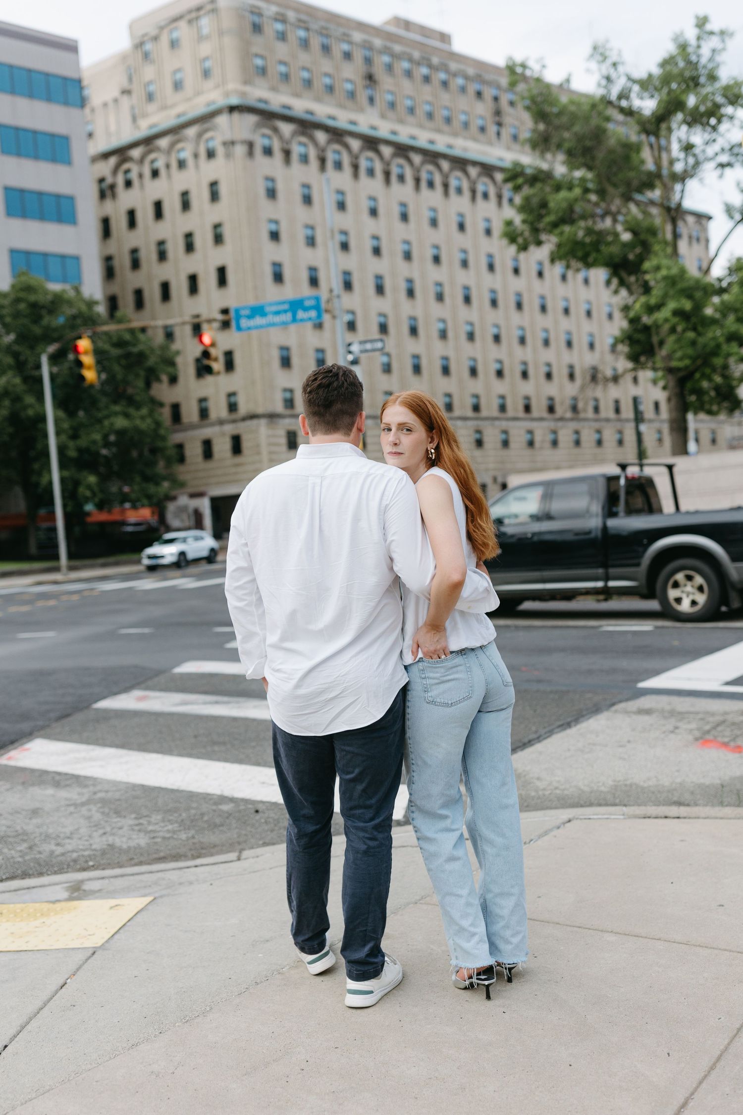 A couple stands together at a crosswalk in an urban setting with tall buildings in the background.