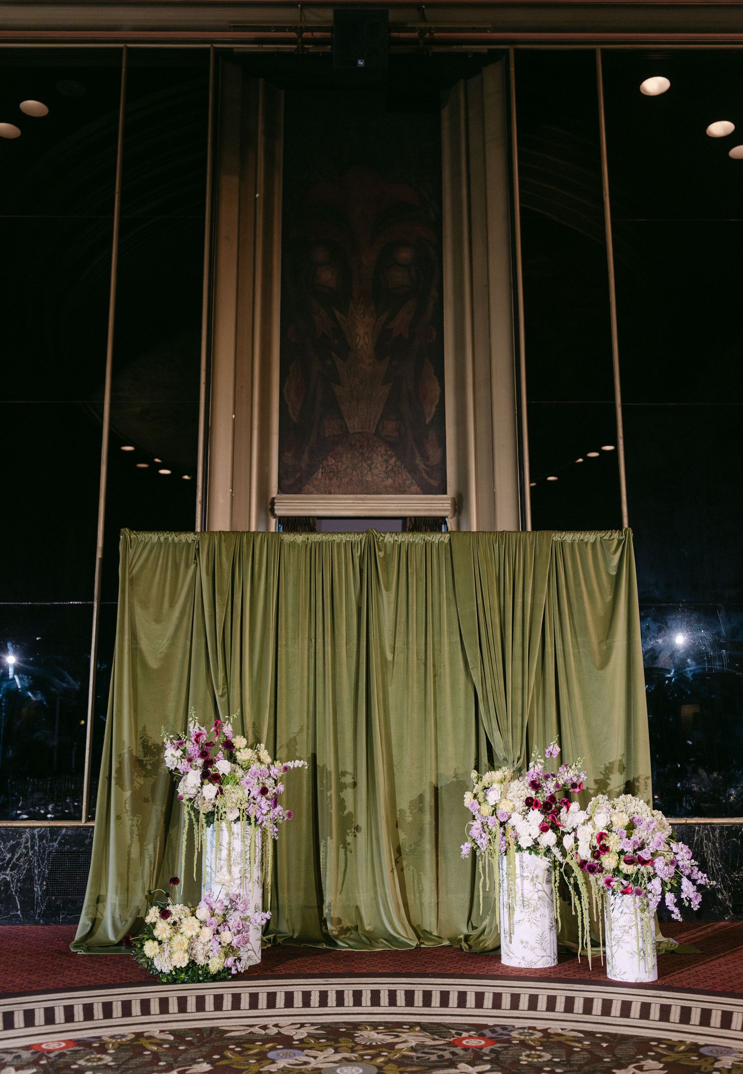 Large elegant wedding venue with green curtains and floral arrangements displayed on surrounding stage area.