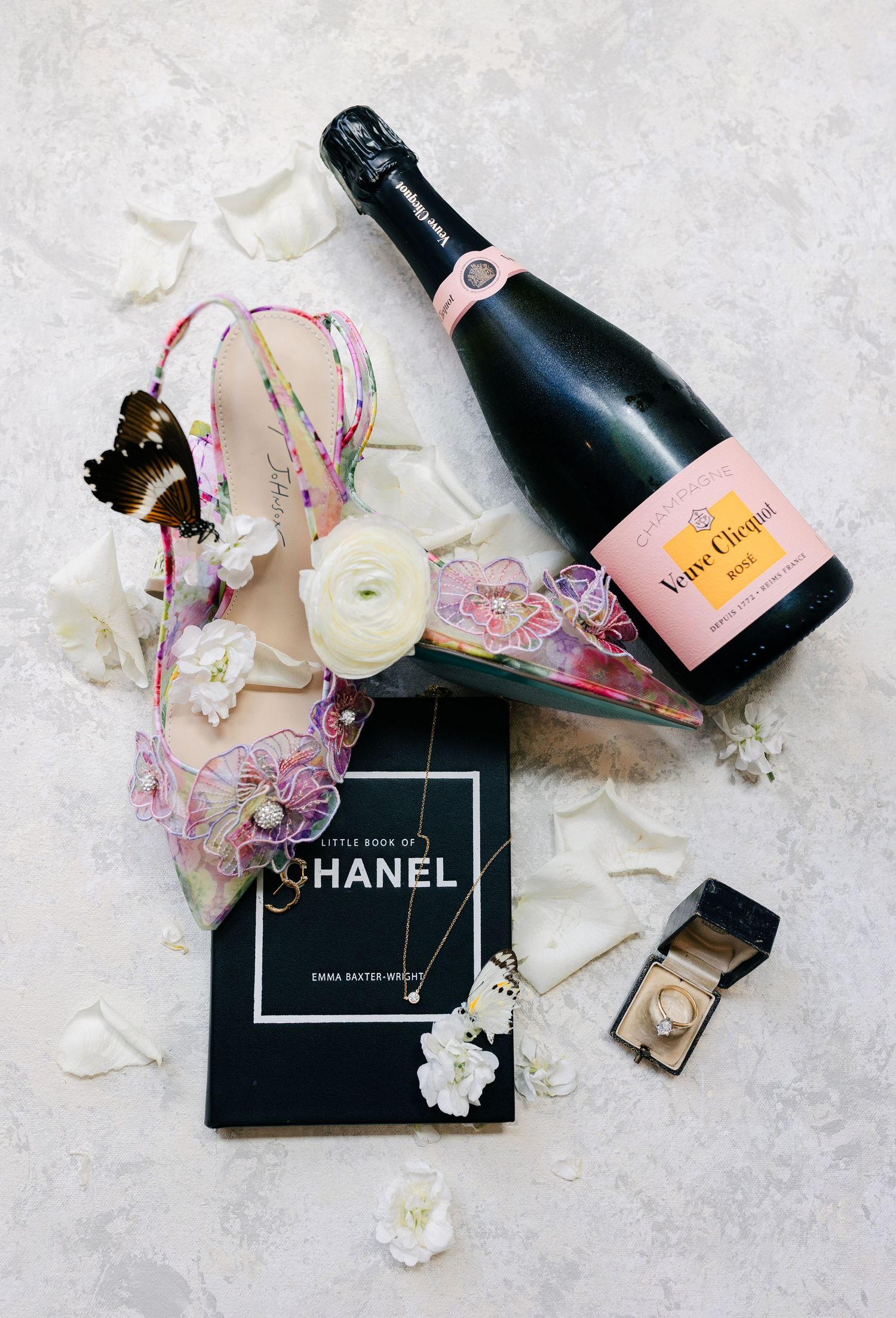 Luxury flatlay with Veuve Clicquot champagne bottle, Chanel book, scattered white flowers and elegant accessories.