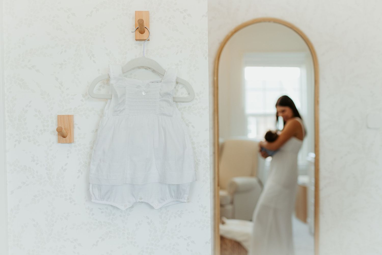 A white christening gown hangs on a hanger next to an arched mirror in a room.