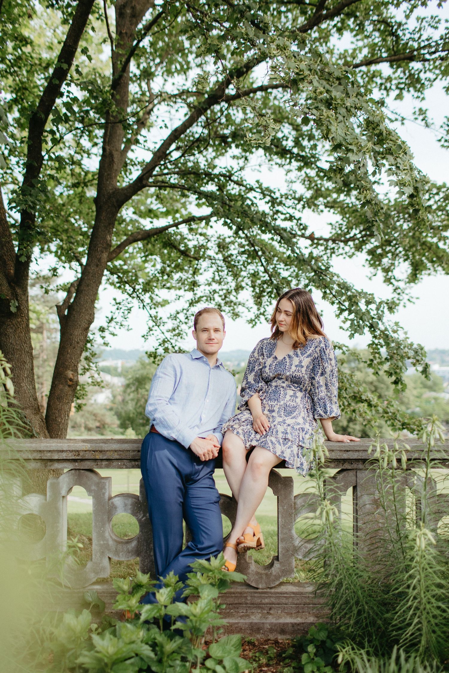 Couple sitting on stone wall surrounded by lush green trees.
